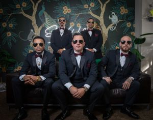 Groomsmen in black suits and sunglasses pose on a couch against a decorative wallpaper background.