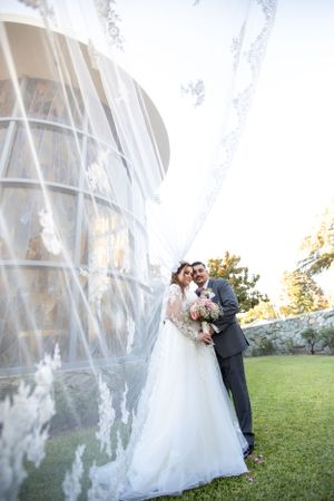 A wedding couple poses outside a modern glass building with flowing veil in natural sunlight.