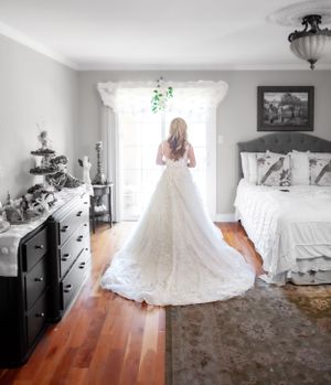 A wedding dress with a long train is photographed from behind in a sunlit bedroom.