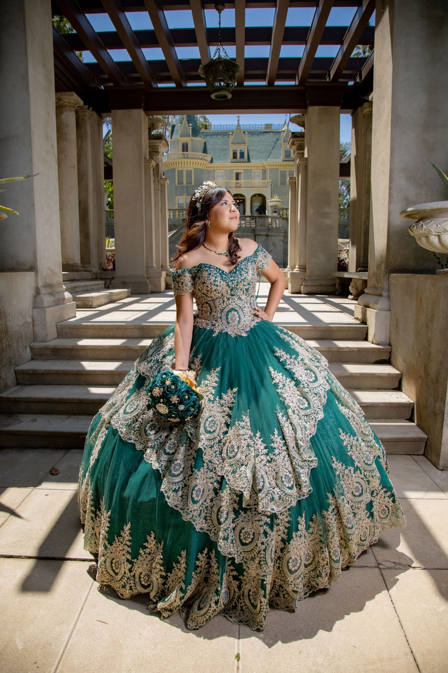 Young woman in an elegant teal and gold quinceañera gown poses under a grand colonnade.