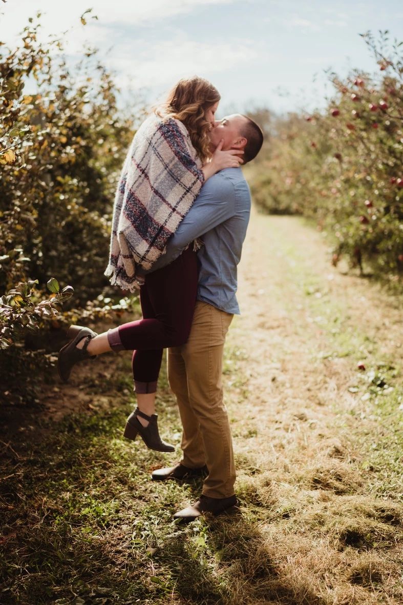 A couple embracing in an orchard with one person lifted off the ground in a romantic moment.