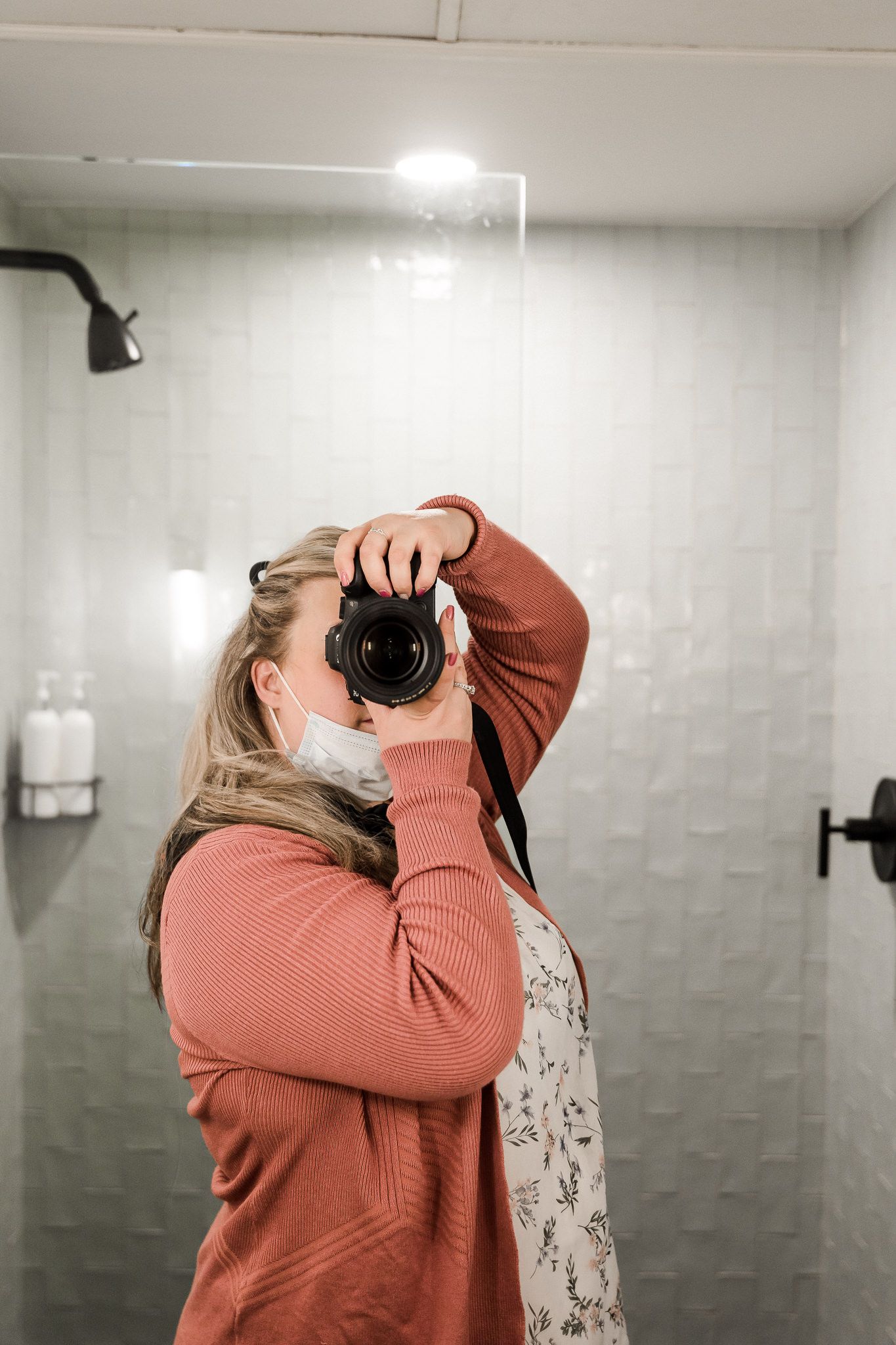 Photographer in coral sweater taking pictures with camera in bathroom setting.