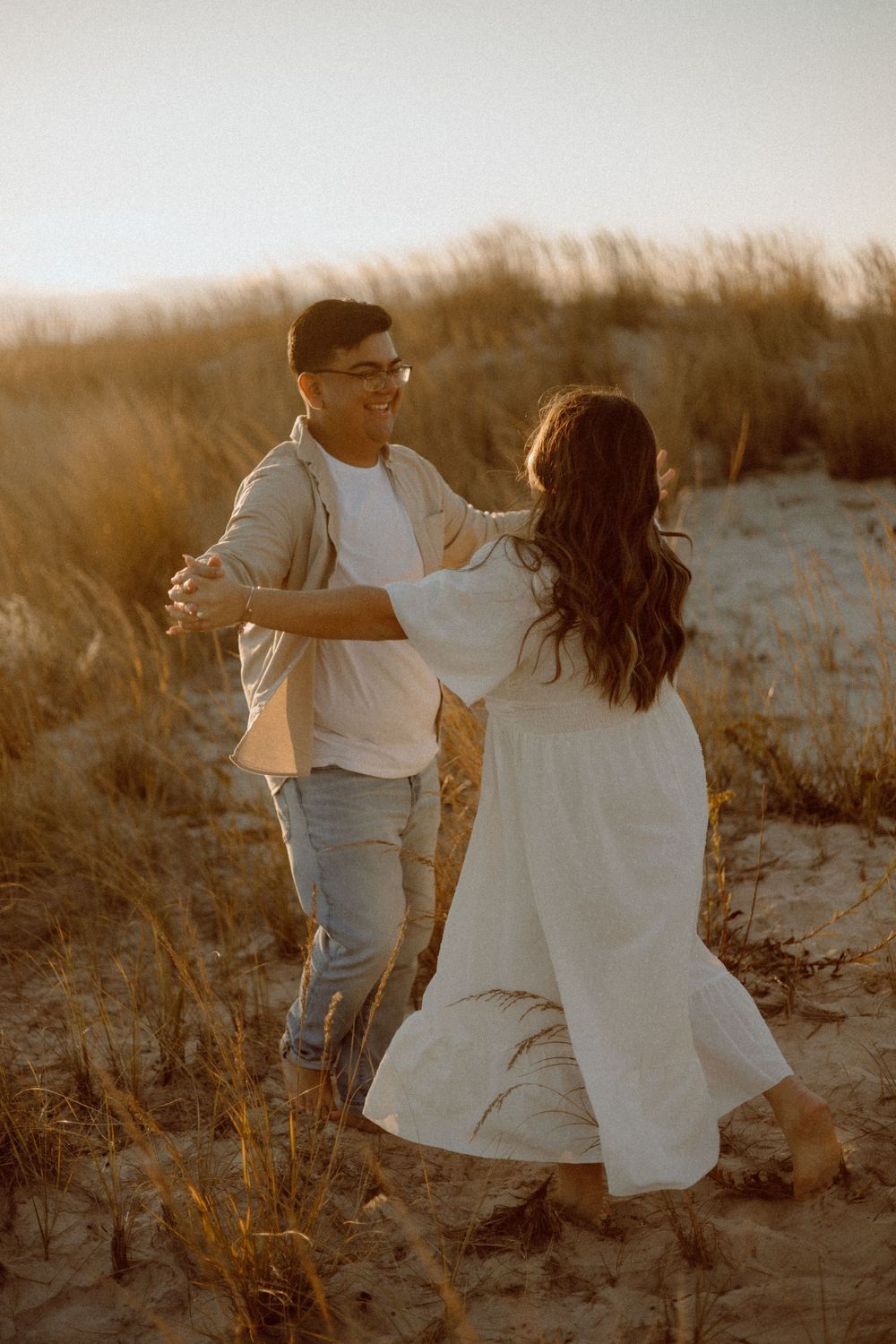 A romantic couple in white clothing dancing in beach dunes during golden hour.
