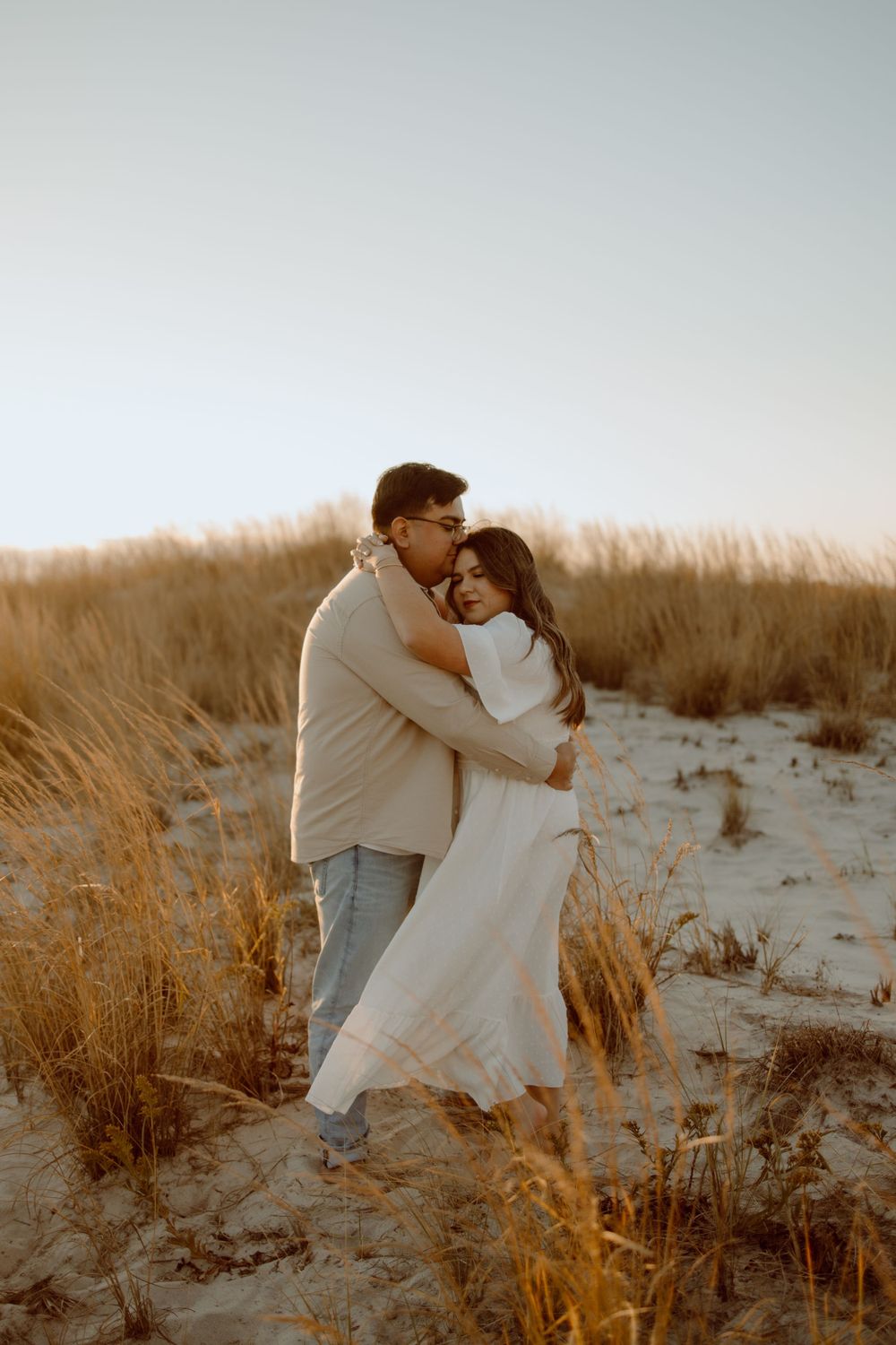 Romantic couple embraces in golden beach grass during sunset photoshoot.