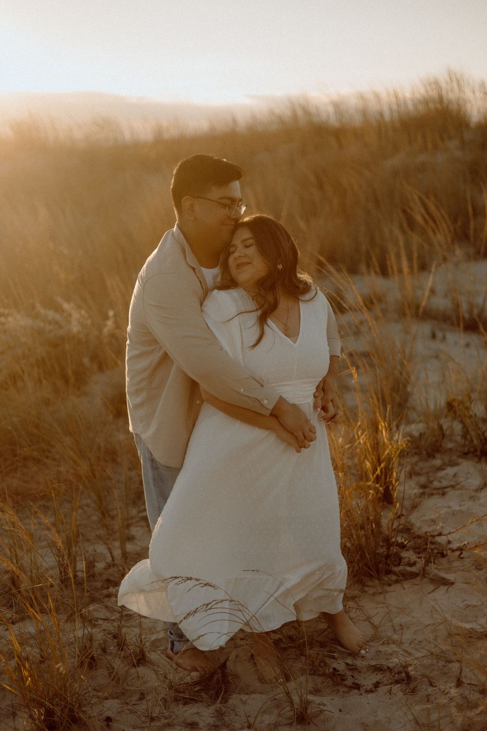 A tender embrace between two people wearing white outfits among beach grass at sunset.
