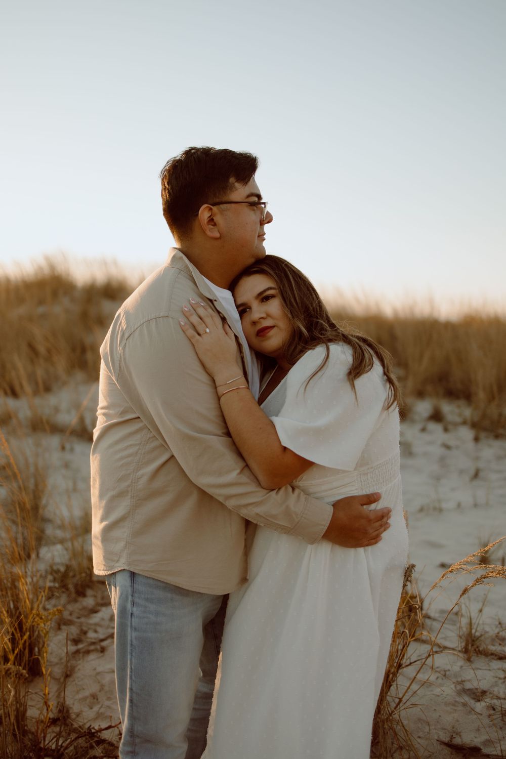 Intimate embrace between couple wearing light colored clothing against coastal dunes.