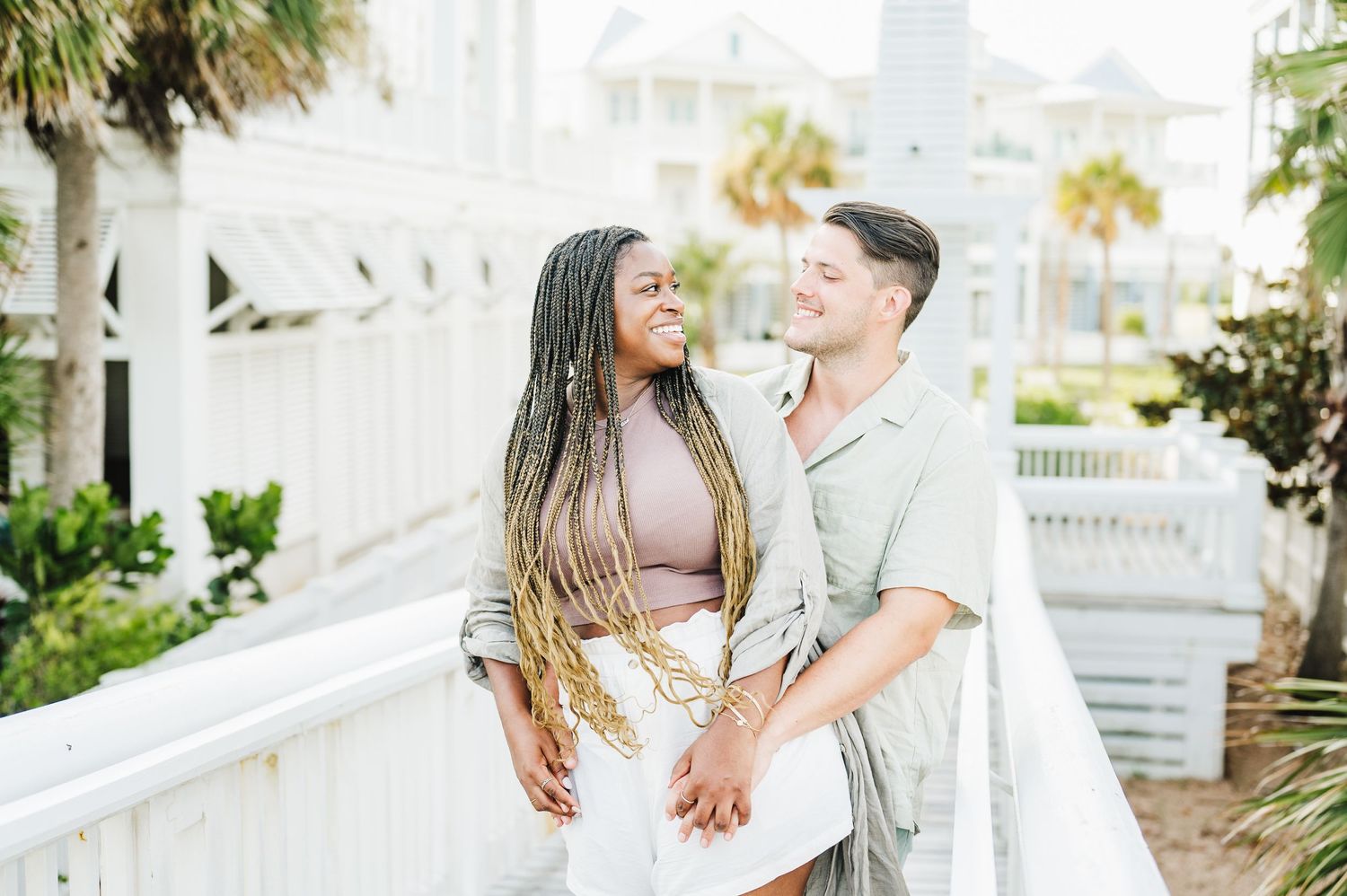 Romantic couple posing on white wooden boardwalk with palm trees and buildings in background.