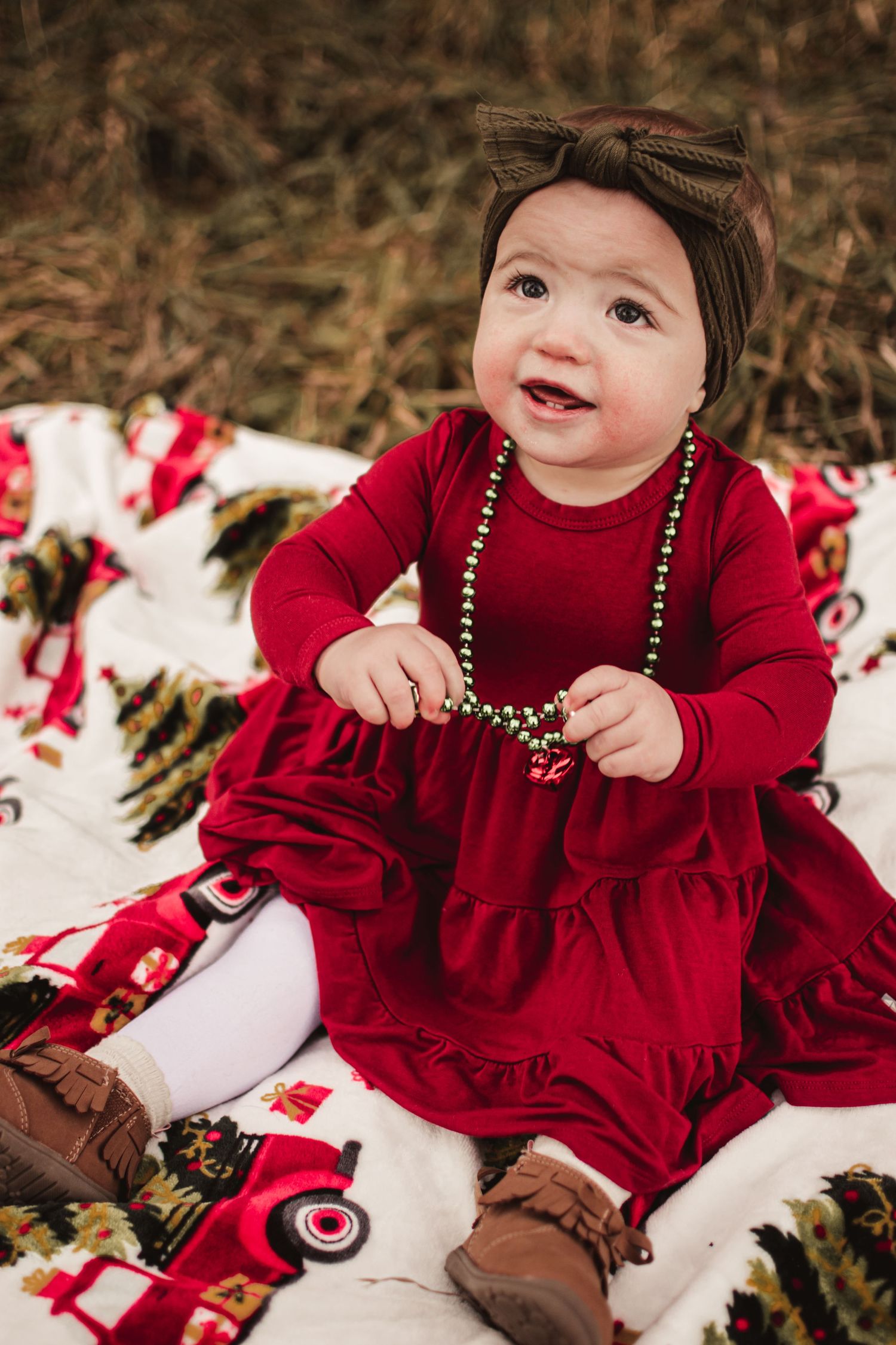 Baby in red dress with beads sits on holiday blanket outdoors smiling.