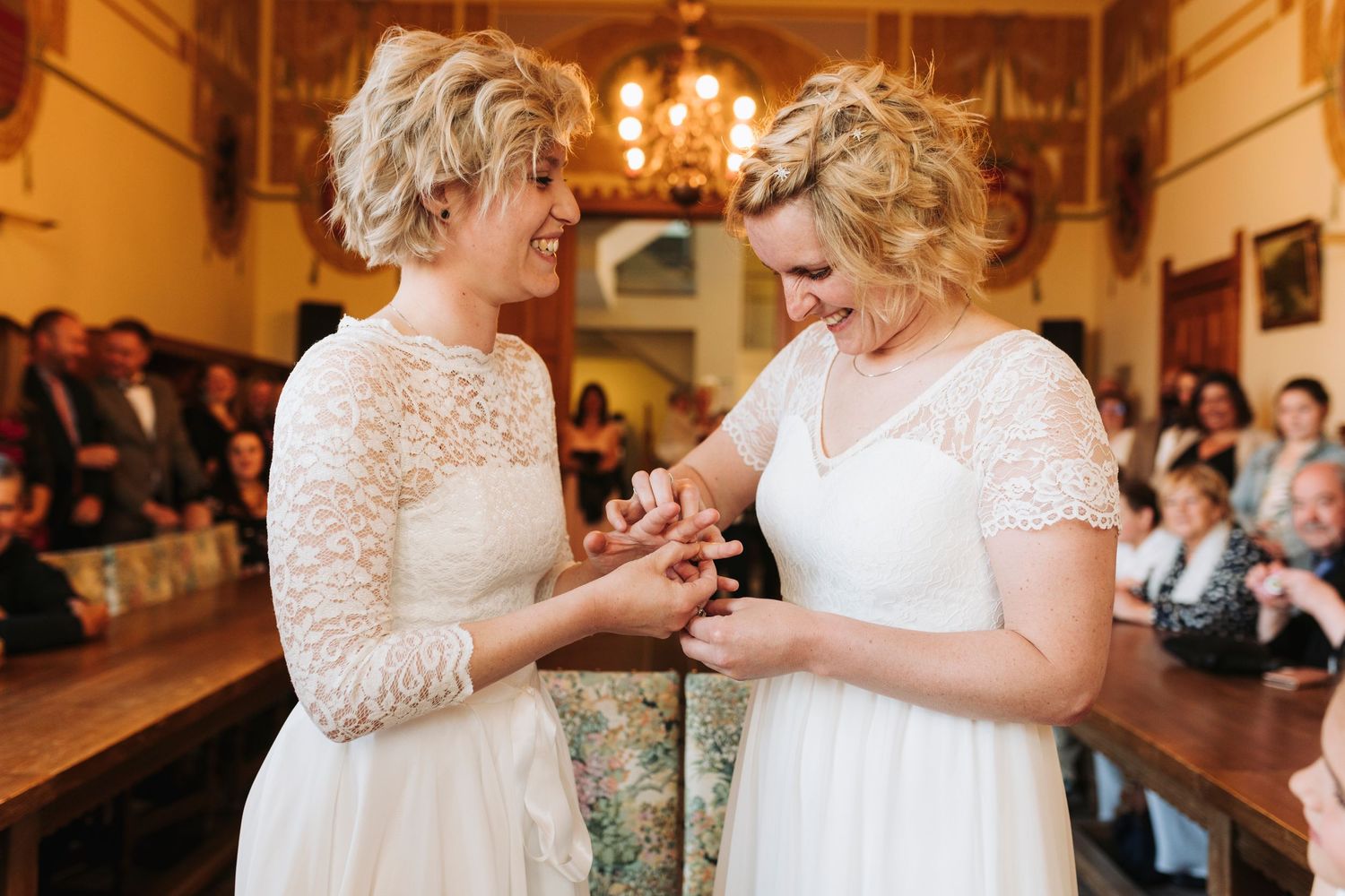 Two people in white lace dresses exchanging rings during a ceremony.
