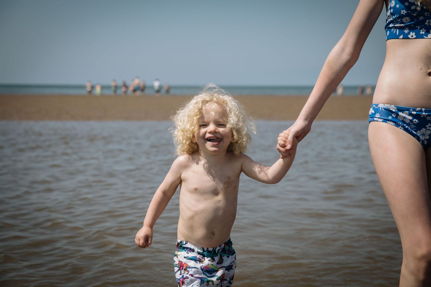 Child plays in shallow ocean water while holding an adult's hand on a sunny beach day.