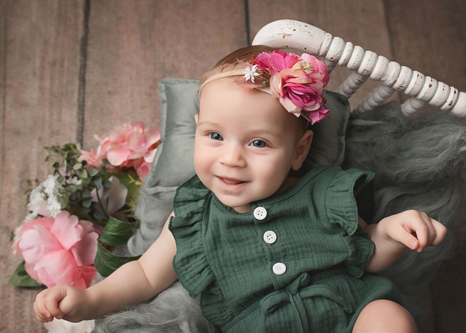 Baby in green ruffled outfit with floral headband poses with pink roses against rustic backdrop.