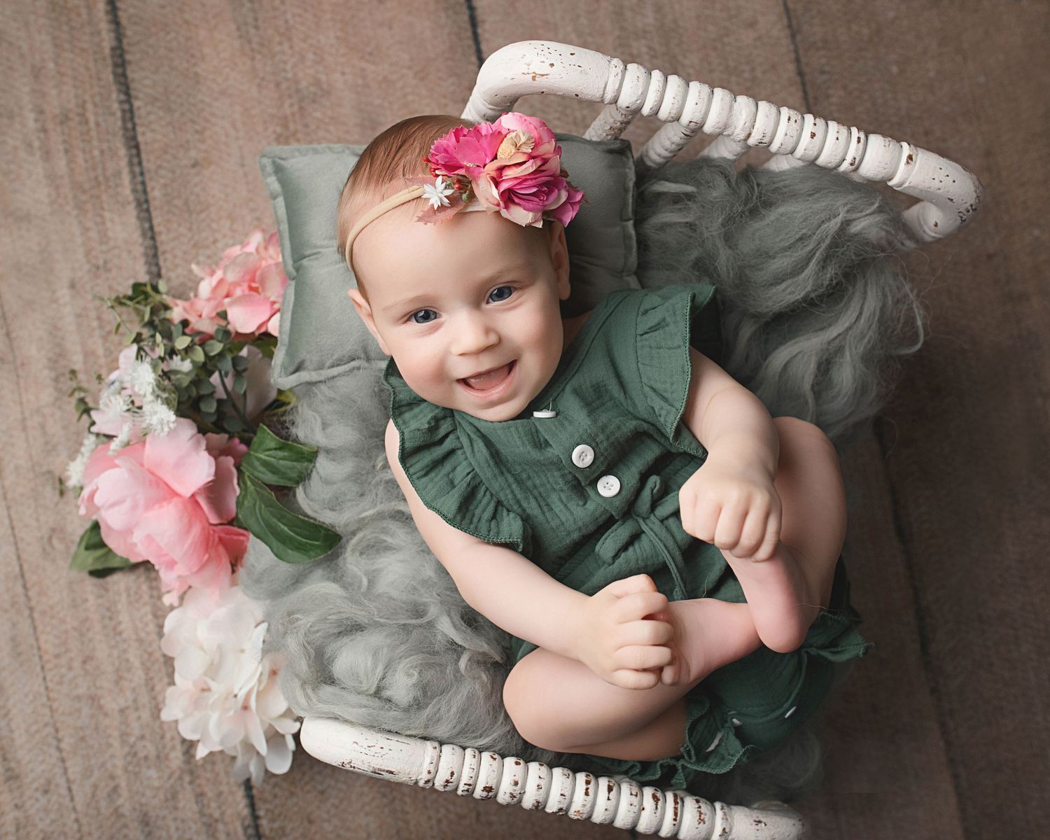 Baby wearing green outfit and pink flower headband poses in white vintage basket with floral decorations.