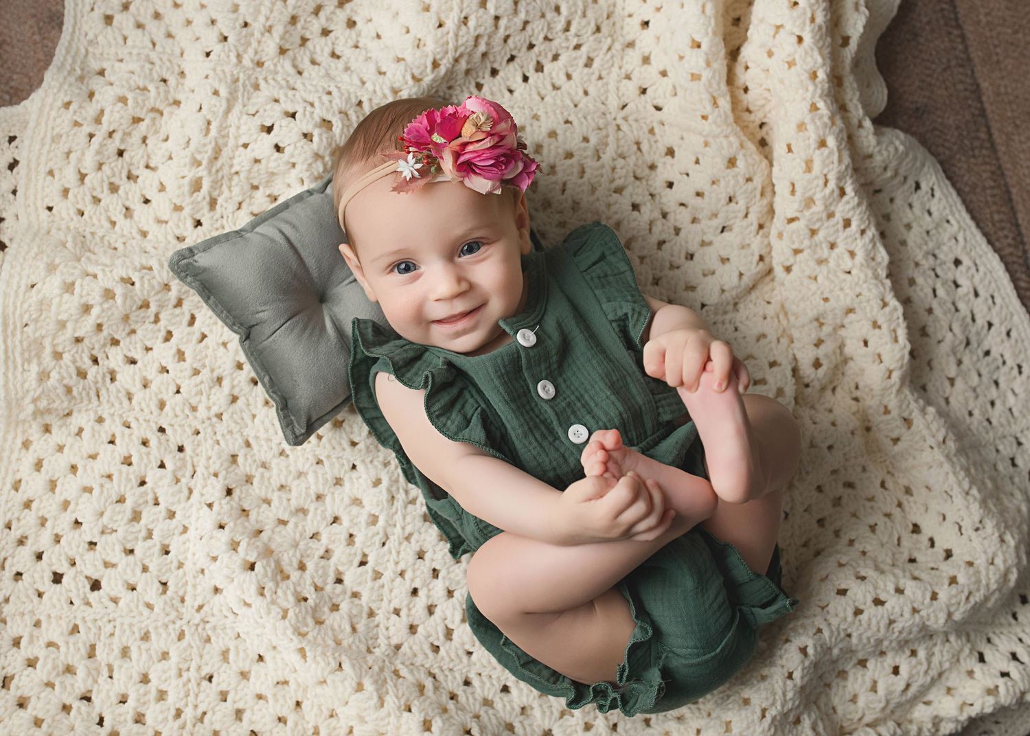 Baby in green romper lying on cream textured blanket with small grey pillow.