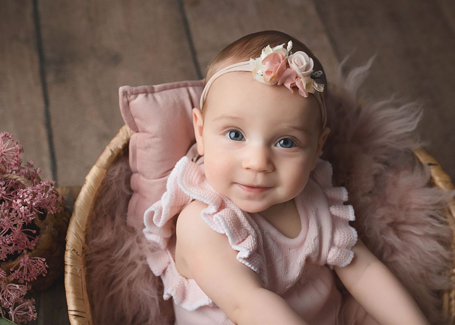 Baby in pink ruffled dress rests in woven basket with soft, vintage-style styling.