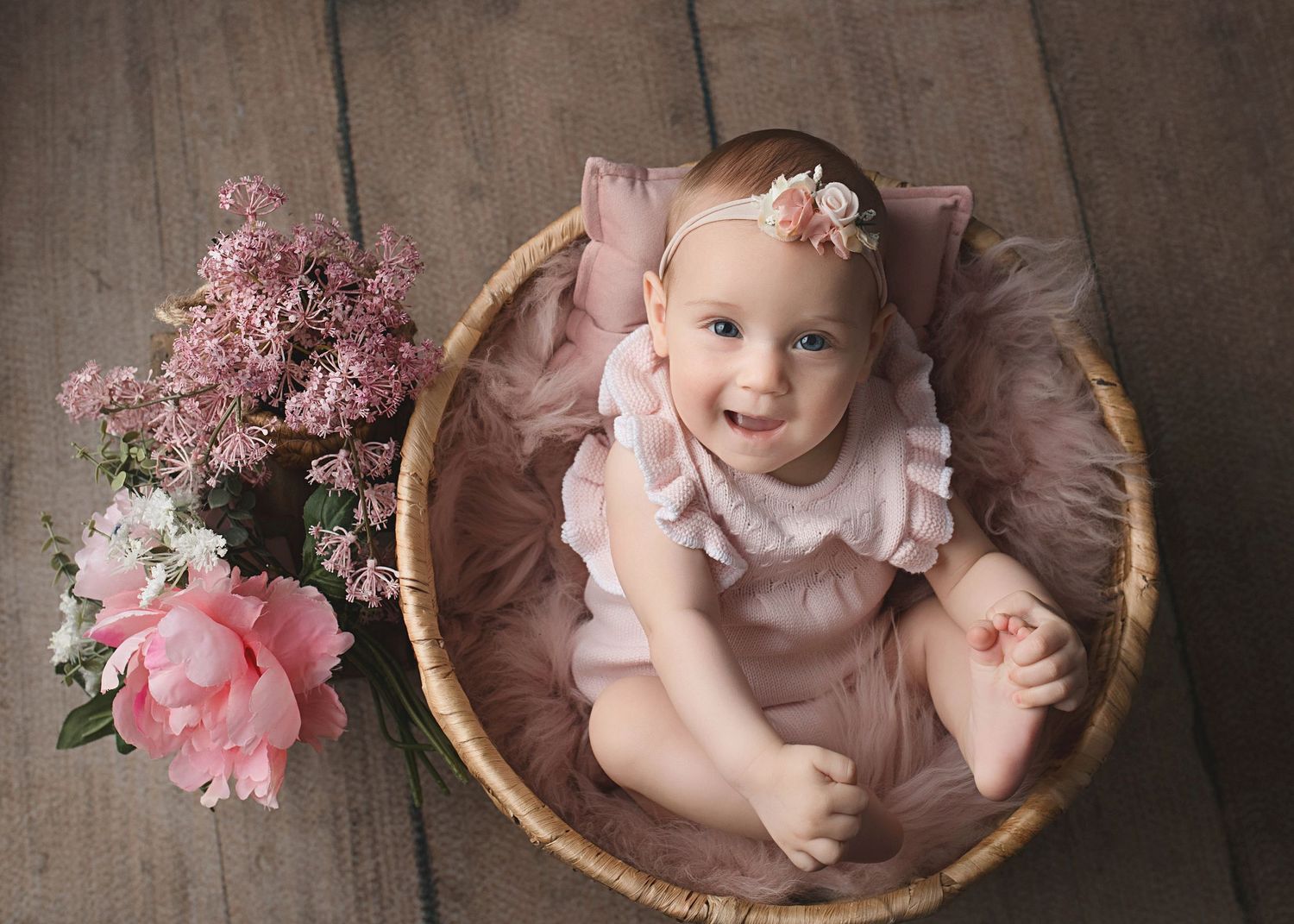 Baby in pink dress posed in basket with pink florals and rustic wooden floor.