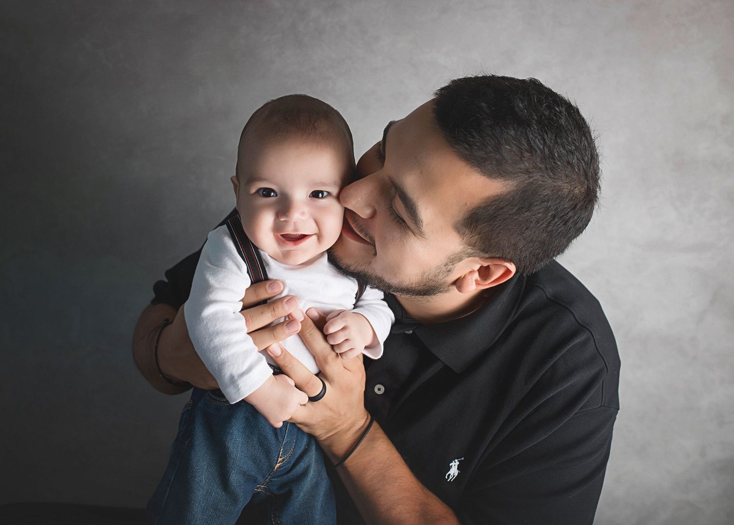 Tender moment captured between parent and baby in casual attire against gray background.