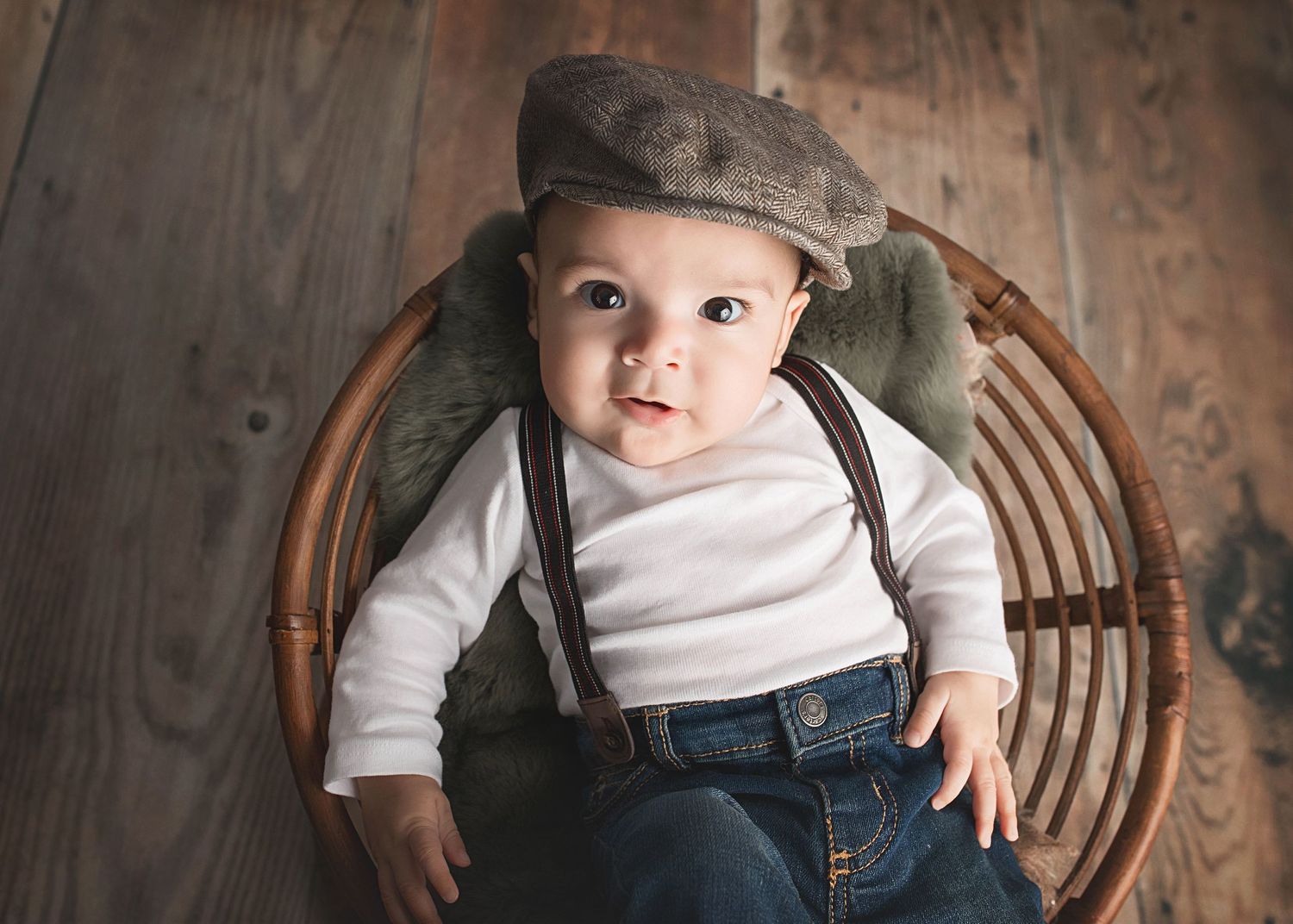 Baby in vintage-inspired outfit with suspenders and cap sits in wicker chair.