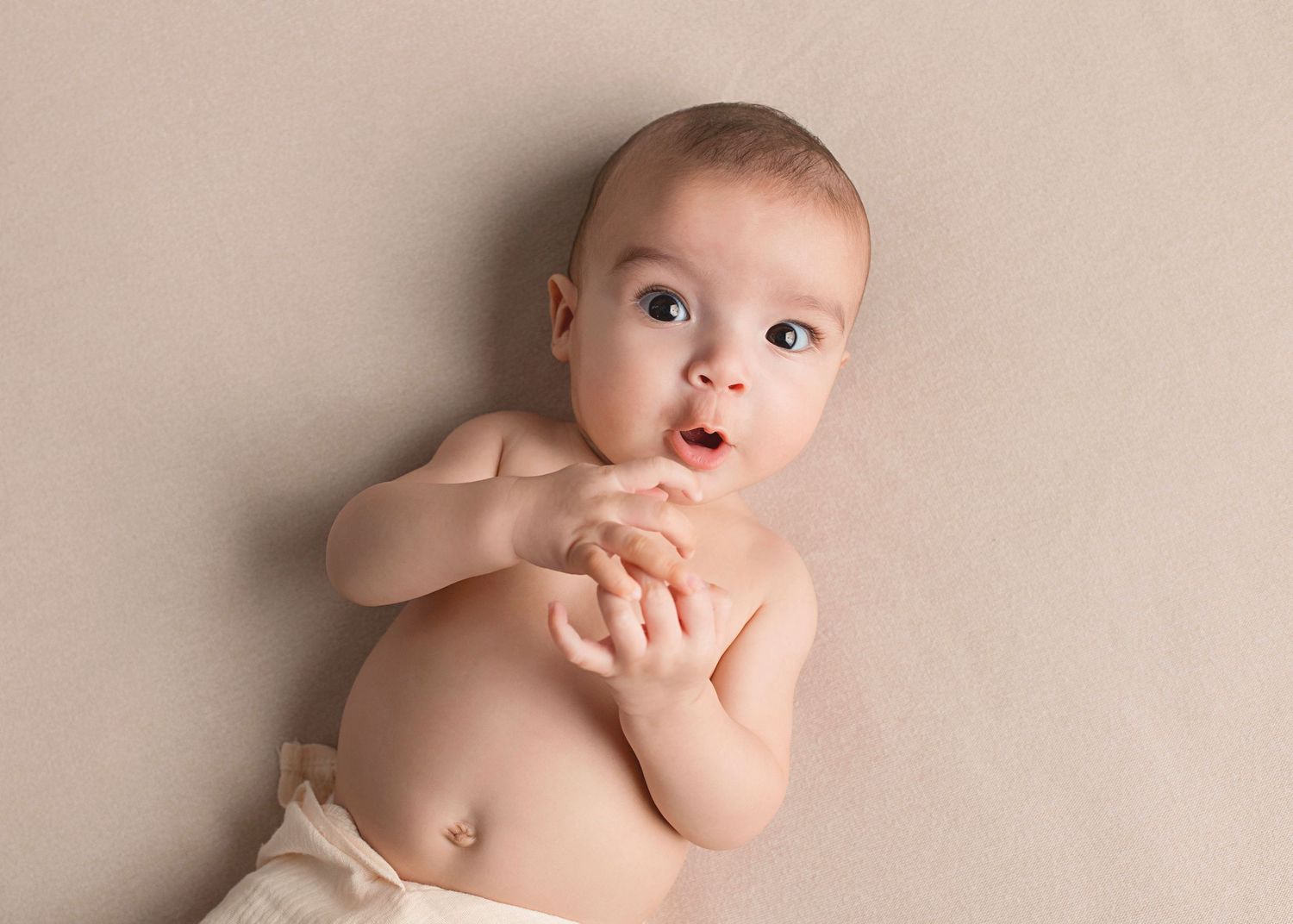 A baby lying on beige backdrop clasps their hands together while gazing upward with wide eyes.