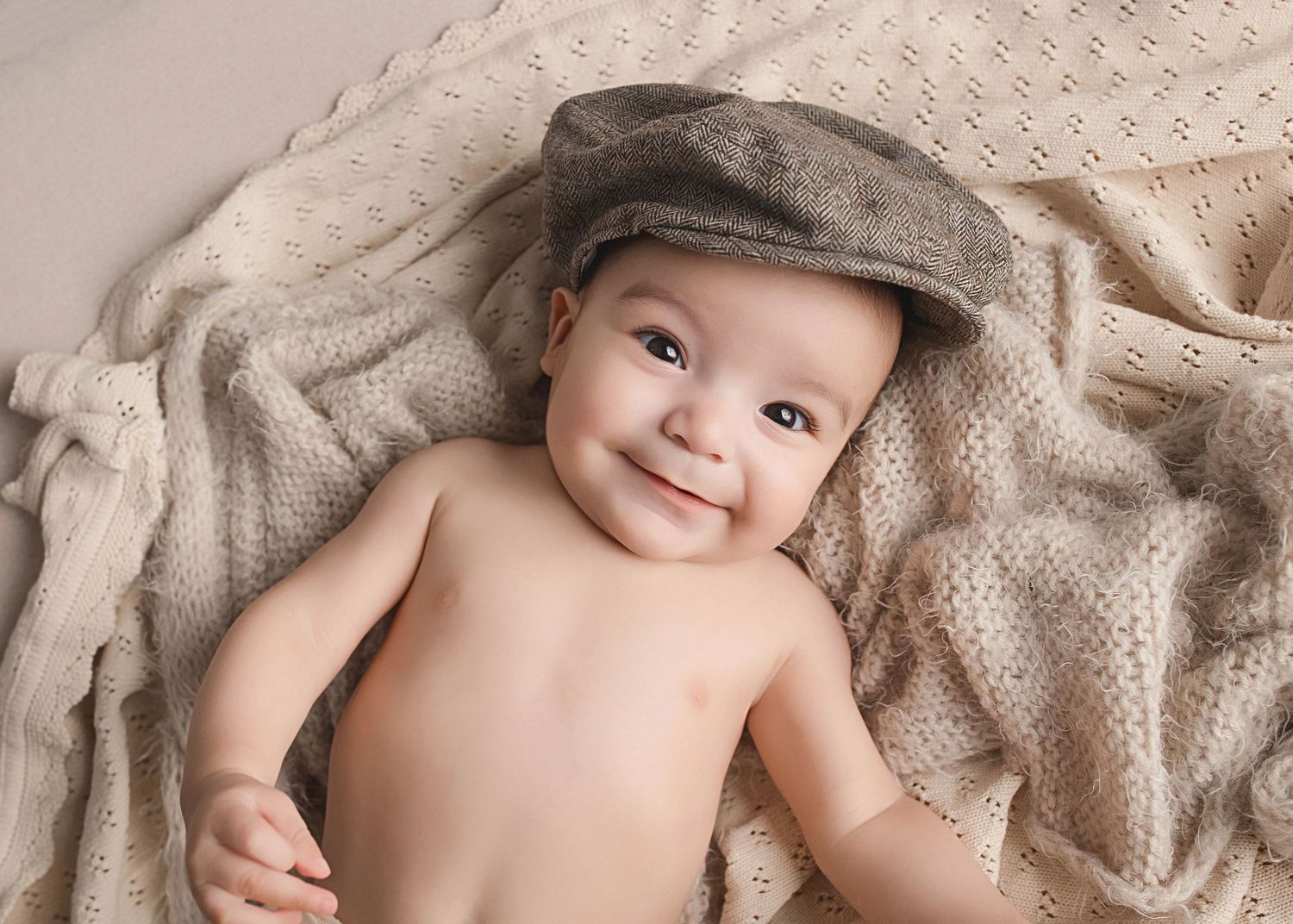A baby wearing a newsboy cap smiles while lying on textured beige fabric.