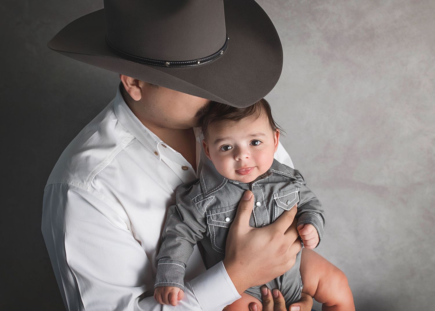 Parent in cowboy hat holds baby wearing grey outfit against neutral studio backdrop.