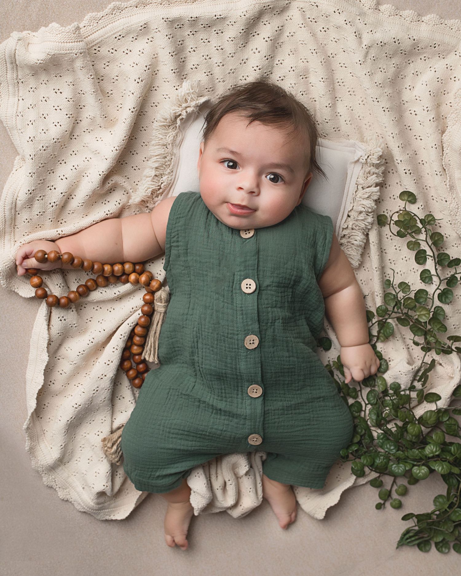 Baby wearing green romper lies on textured cream blanket with wooden beads and eucalyptus accents.