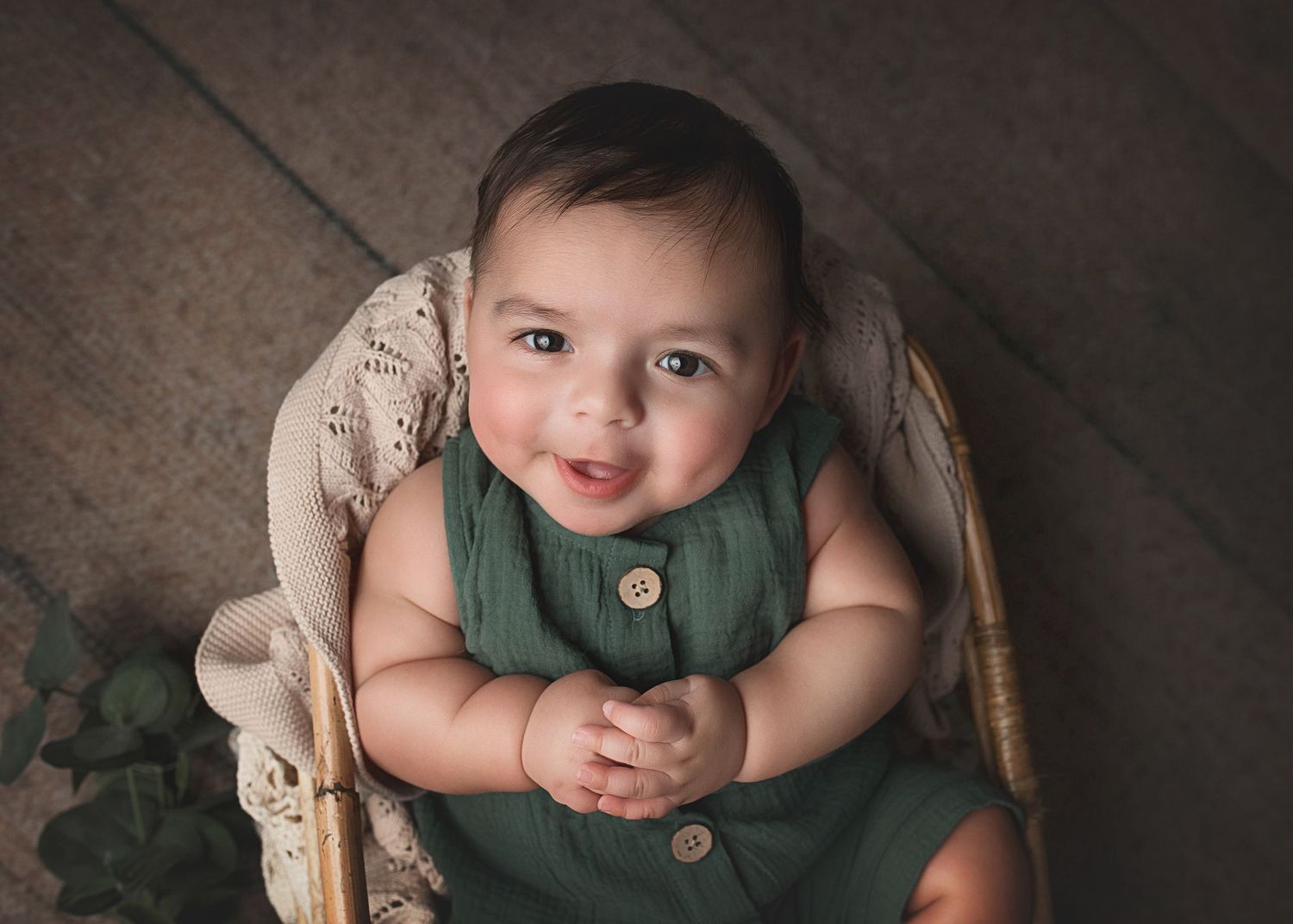 Smiling baby in green button-up romper sits posed in vintage wicker chair.