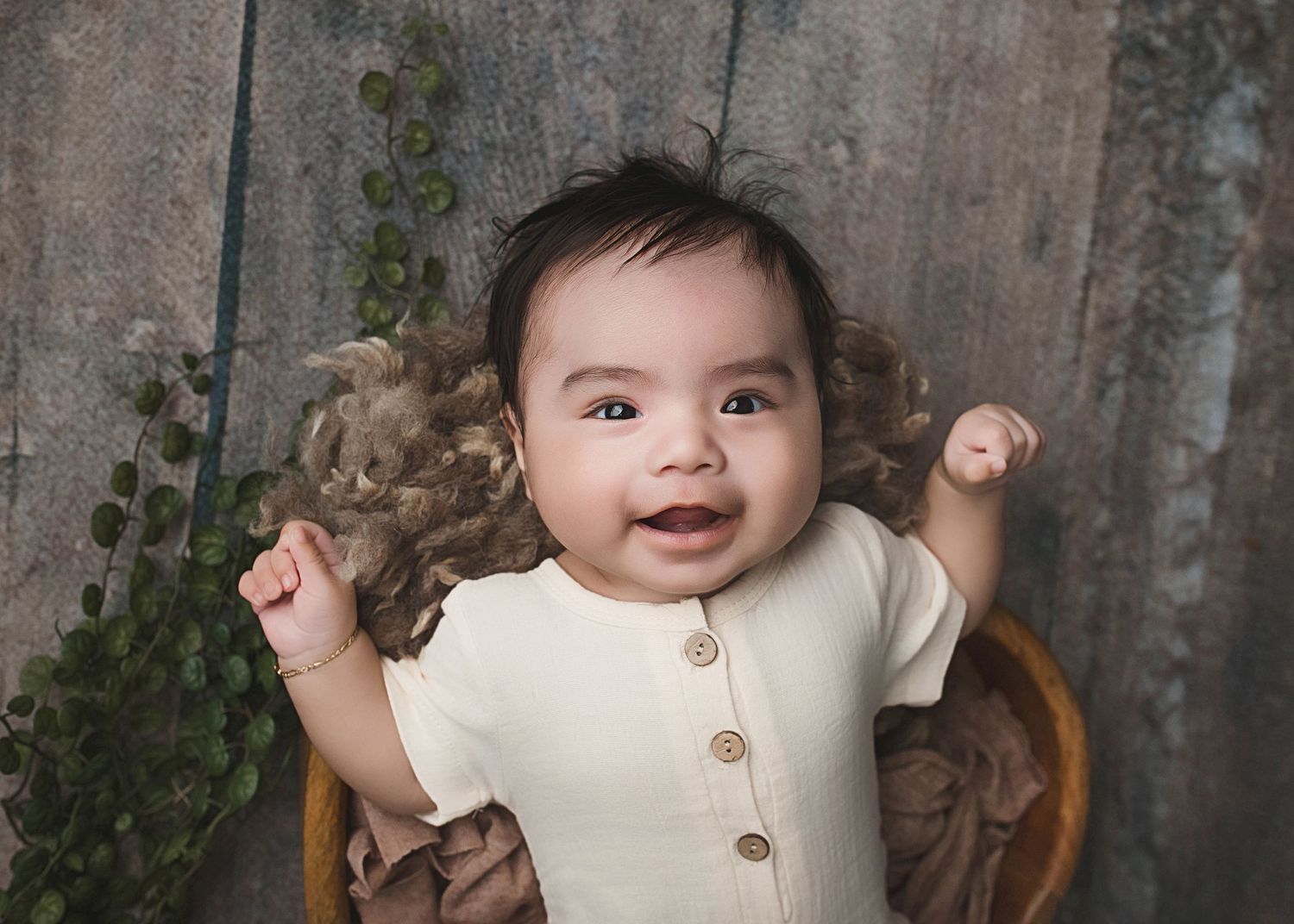 Happy baby in cream-colored button-up outfit smiling while sitting against rustic wooden background.