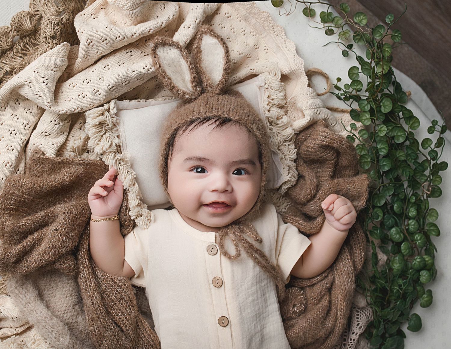 Infant wearing bunny ears and cream romper on soft neutral bedding with ivy decorations.