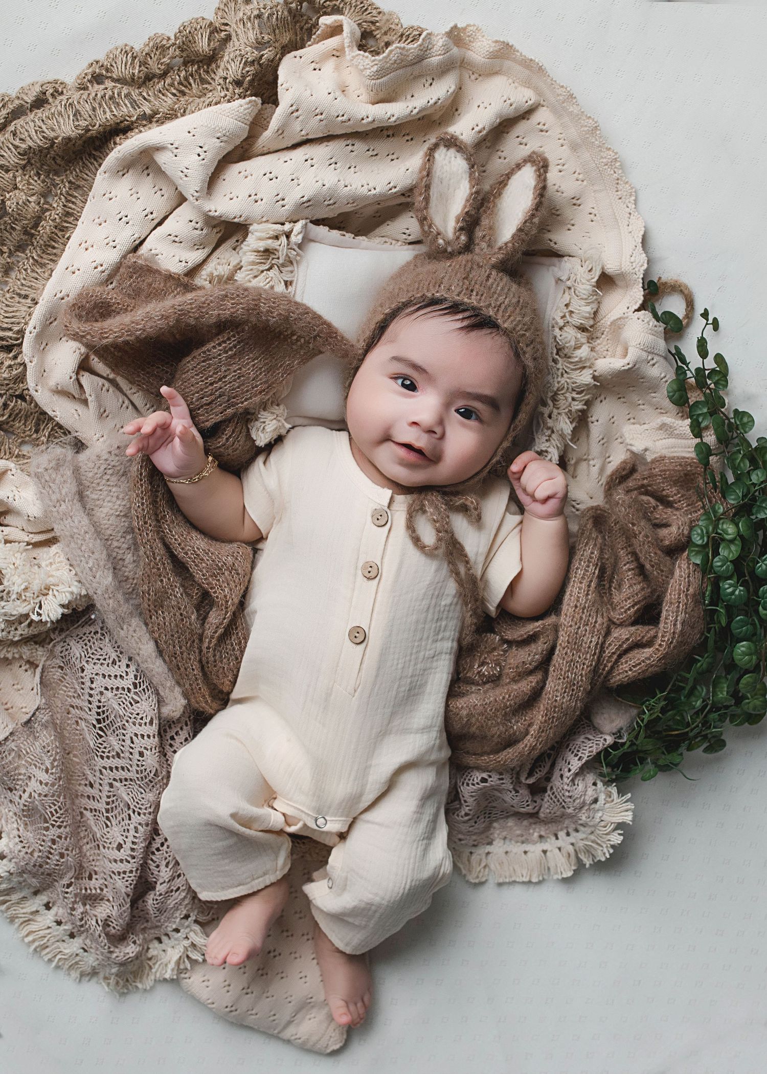 Baby in cream-colored romper lounging on textured beige blanket with decorative greenery.
