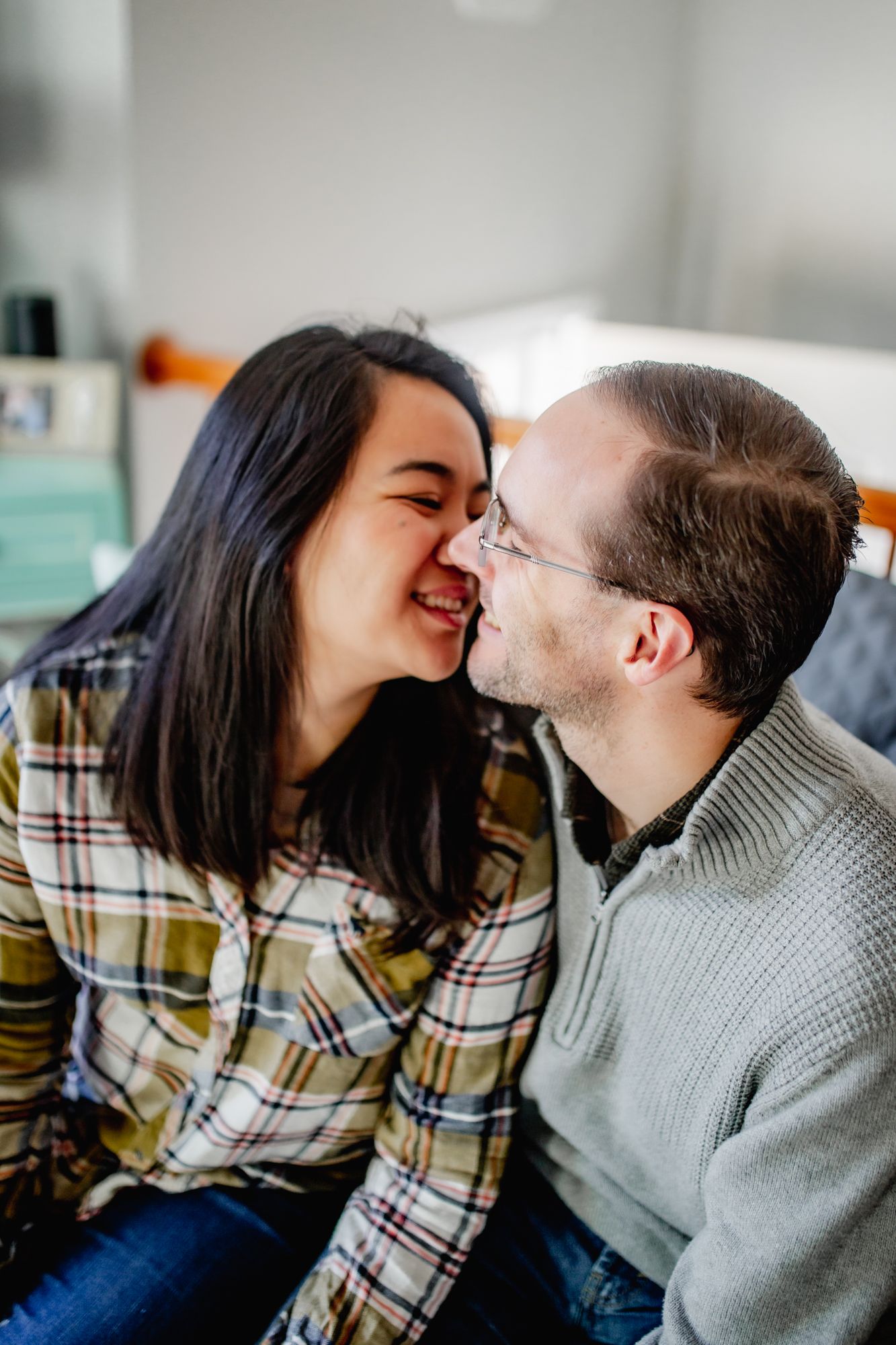 a couple sits together on a couch as they grin and rub noses; she is wearing green flannel and he a grey sweater