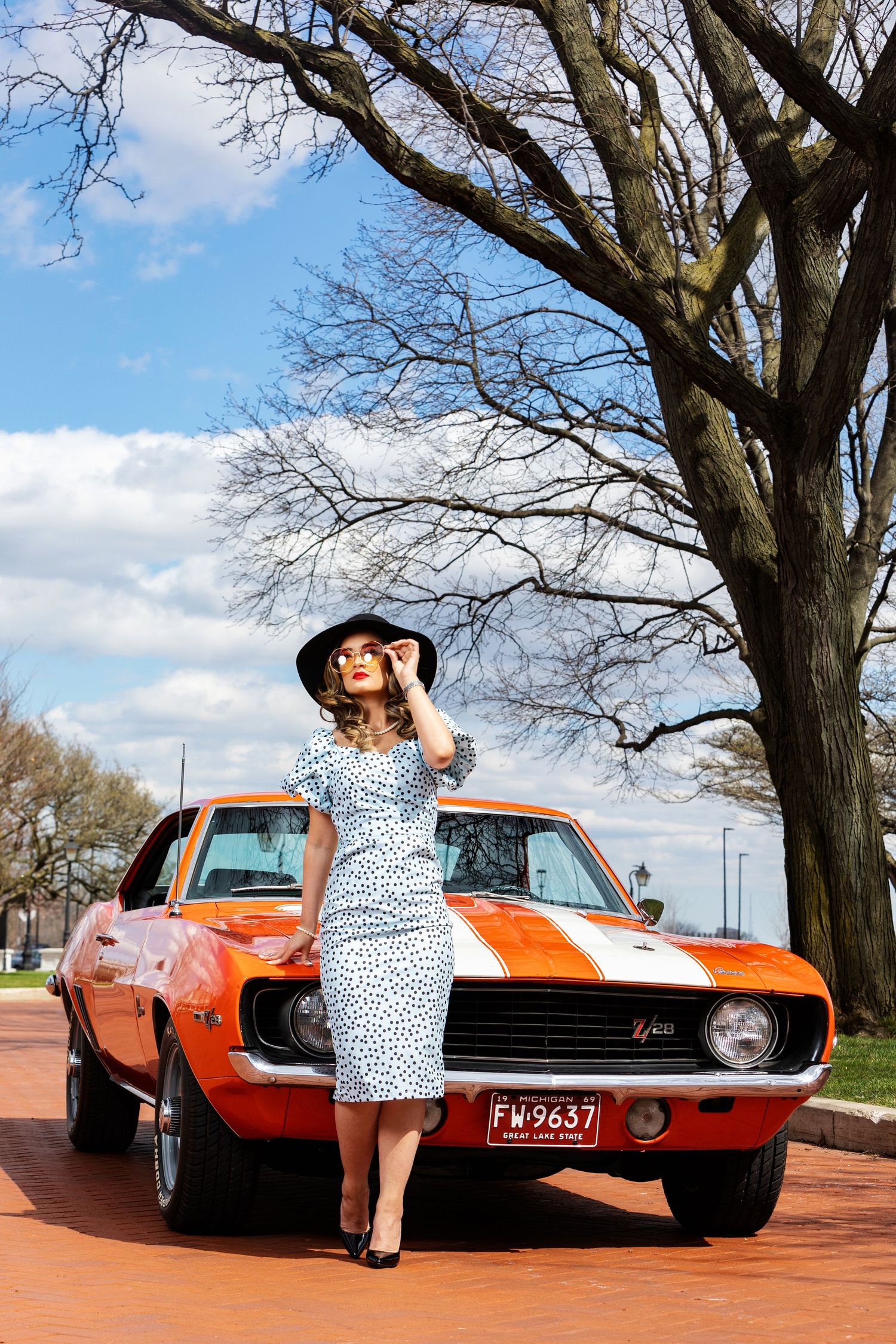 Retro-styled scene with orange classic Camaro and polka dot dress against bare winter tree.