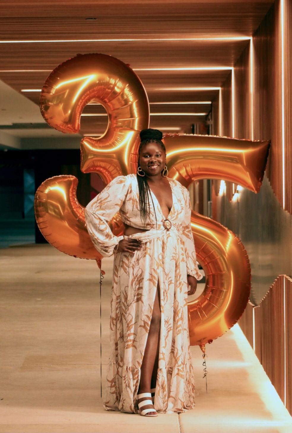 Person in flowing white dress poses with large gold number 35 balloons in an elegant hallway.