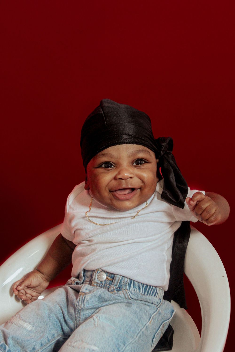 Baby in white shirt and jeans wearing a black durag sits smiling in a chair against a deep red background.