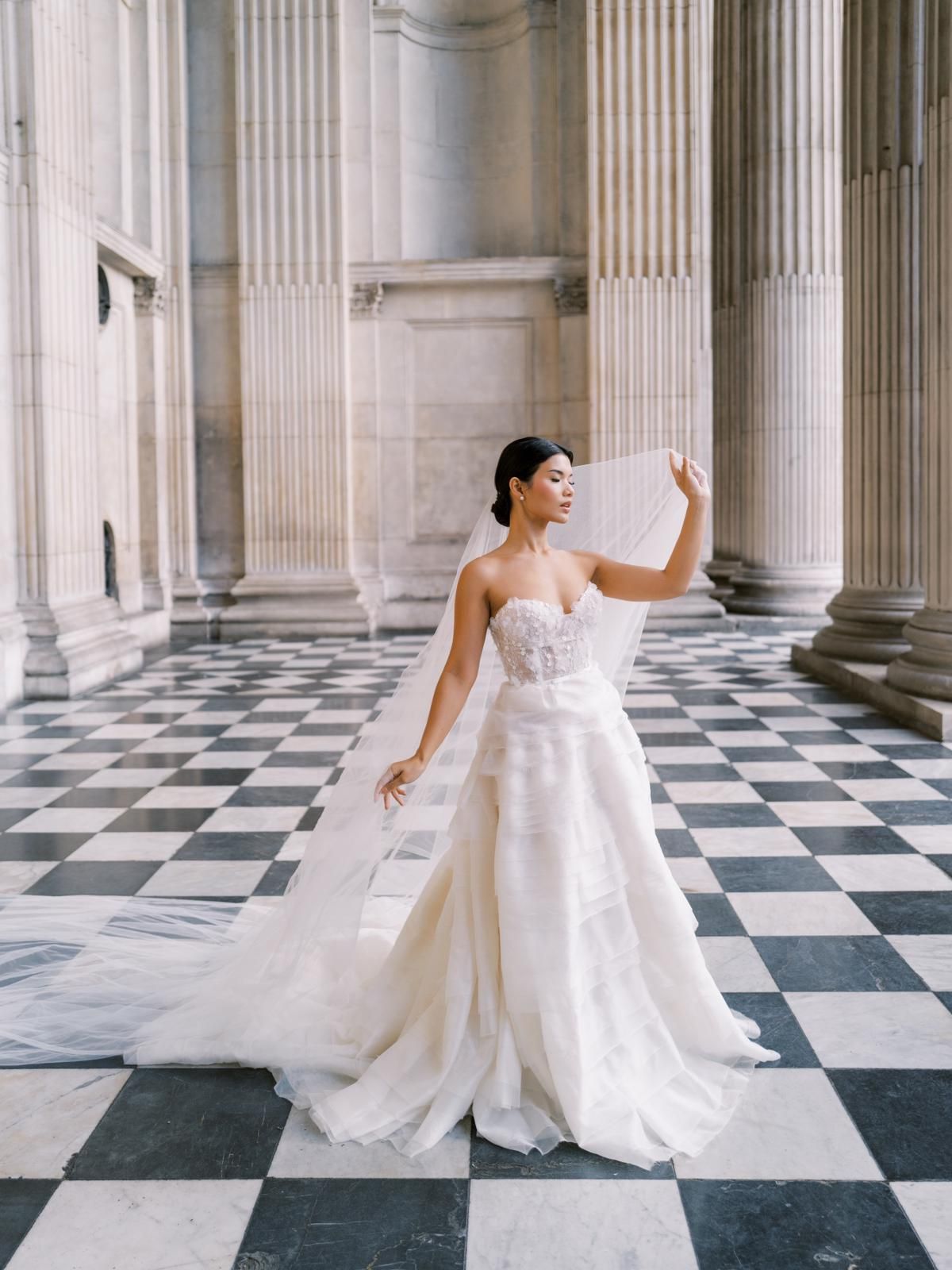 Bride in elegant white gown twirls on black and white checkered floor surrounded by classical columns.