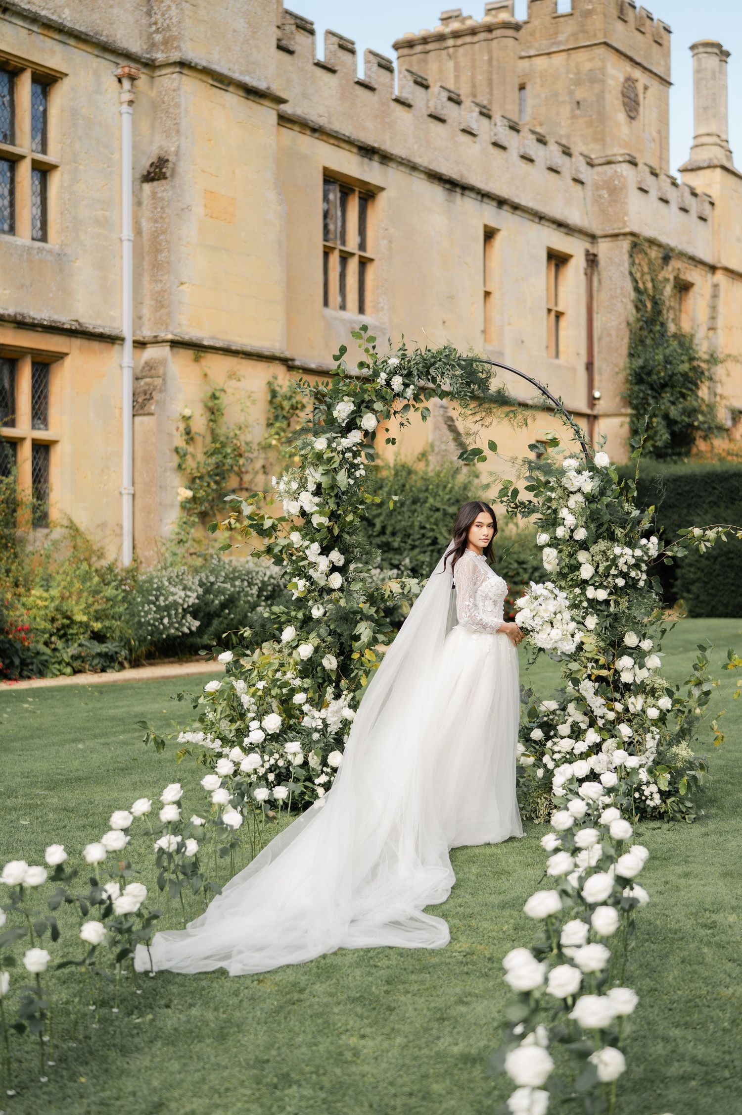 White wedding dress flows elegantly against historic stone castle walls and blooming garden roses.