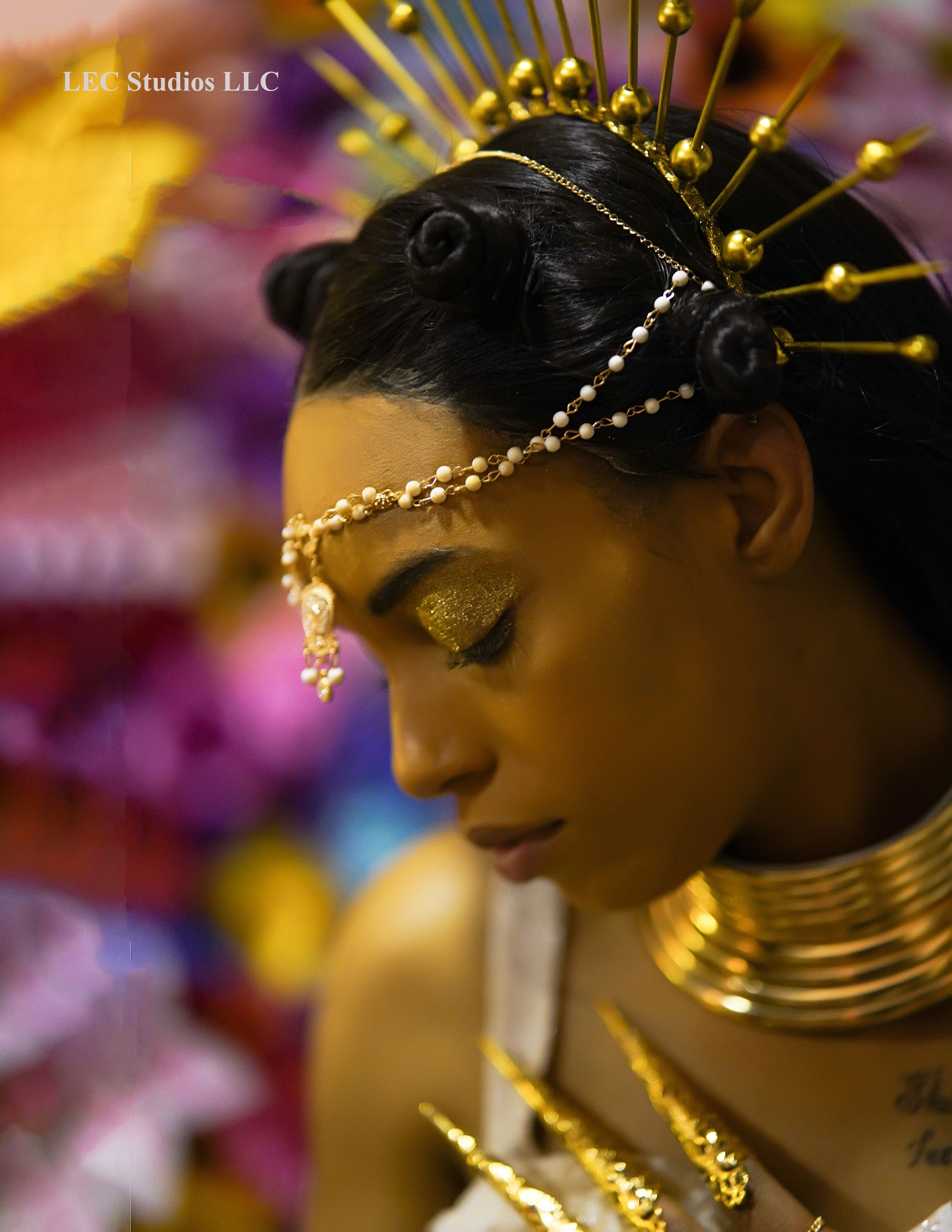 Artistic fashion portrait with ornate gold headpiece and jewelry against colorful floral background.