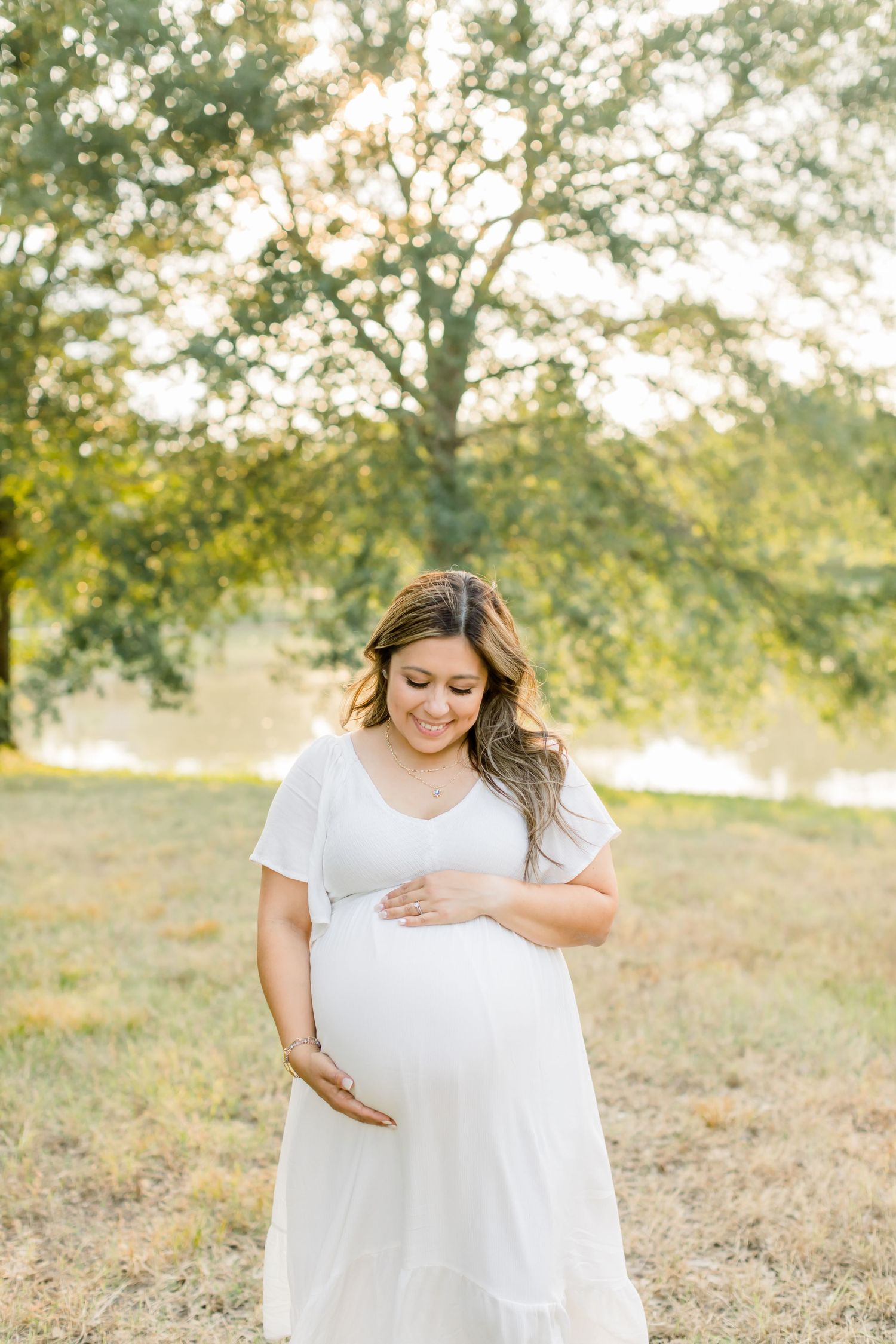 Maternity portrait of woman in white dress tenderly touching baby bump in sunlit field.