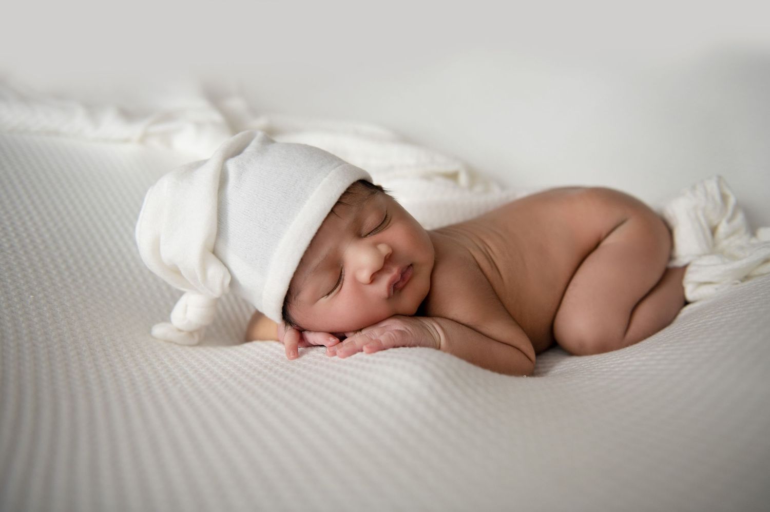 Sleeping newborn wearing white knit hat on textured white background.