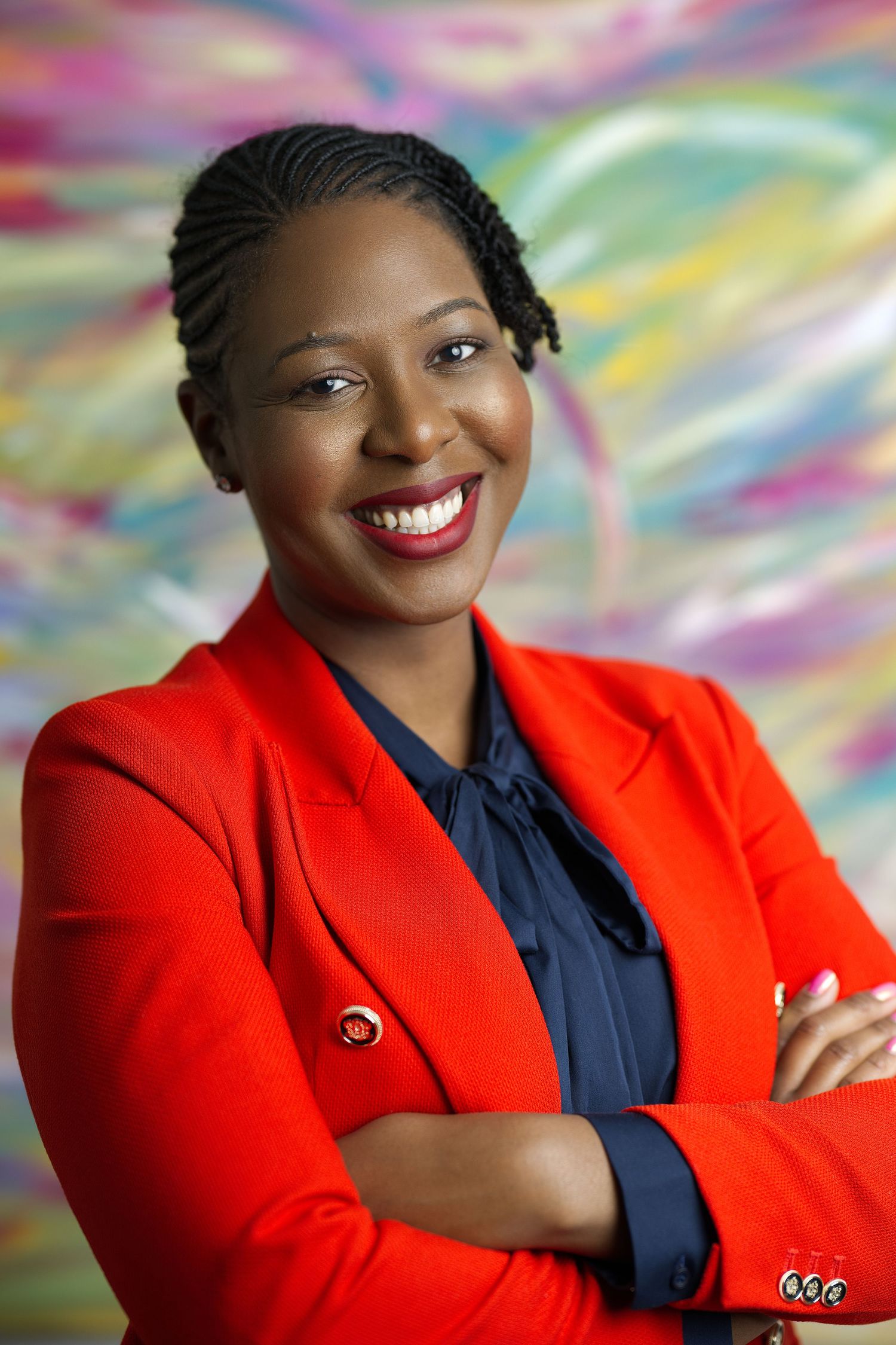 Professional headshot with braided hair wearing a red blazer against a vibrant rainbow backdrop.