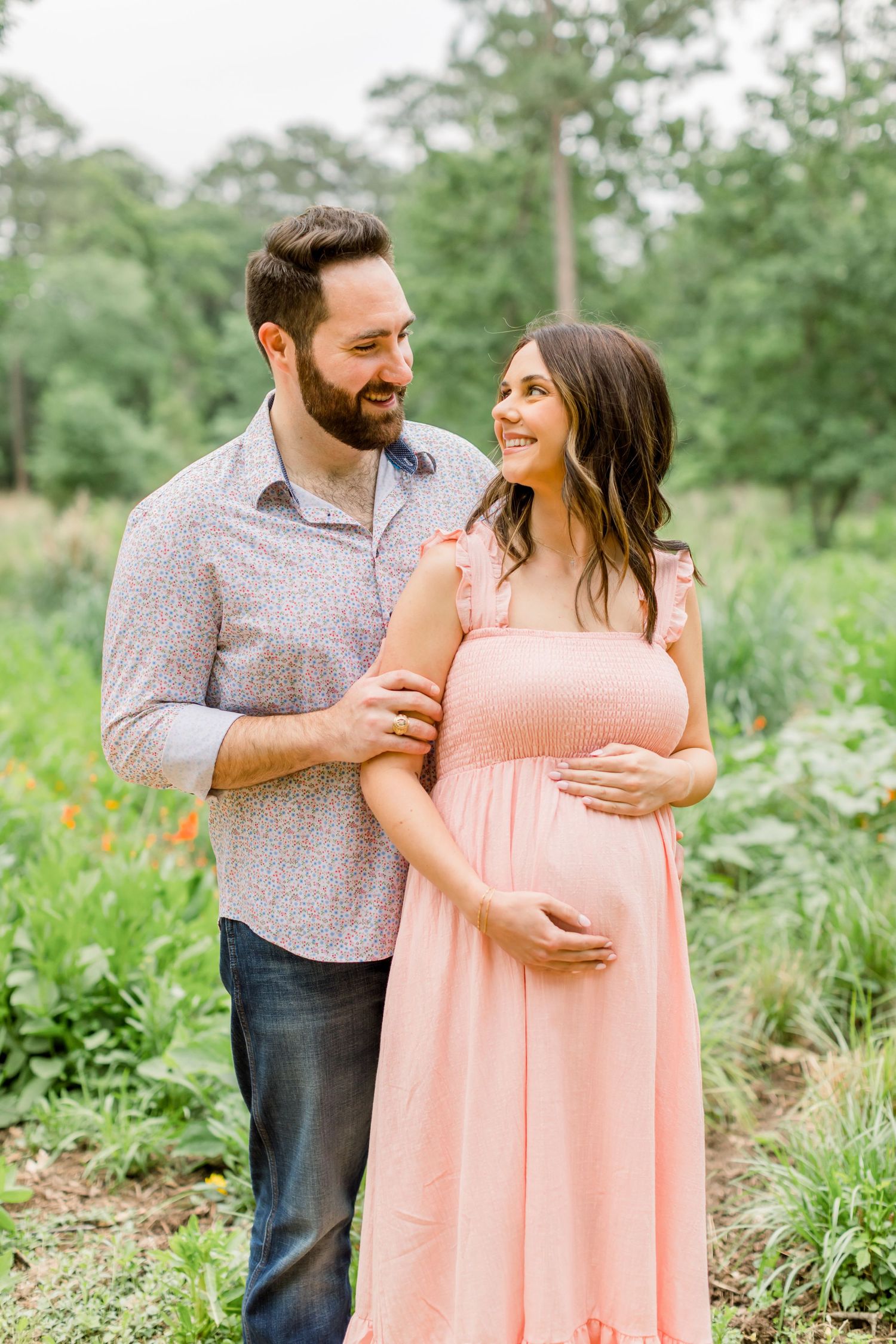 A couple embraces on a nature trail, with the woman wearing a pink maternity dress.