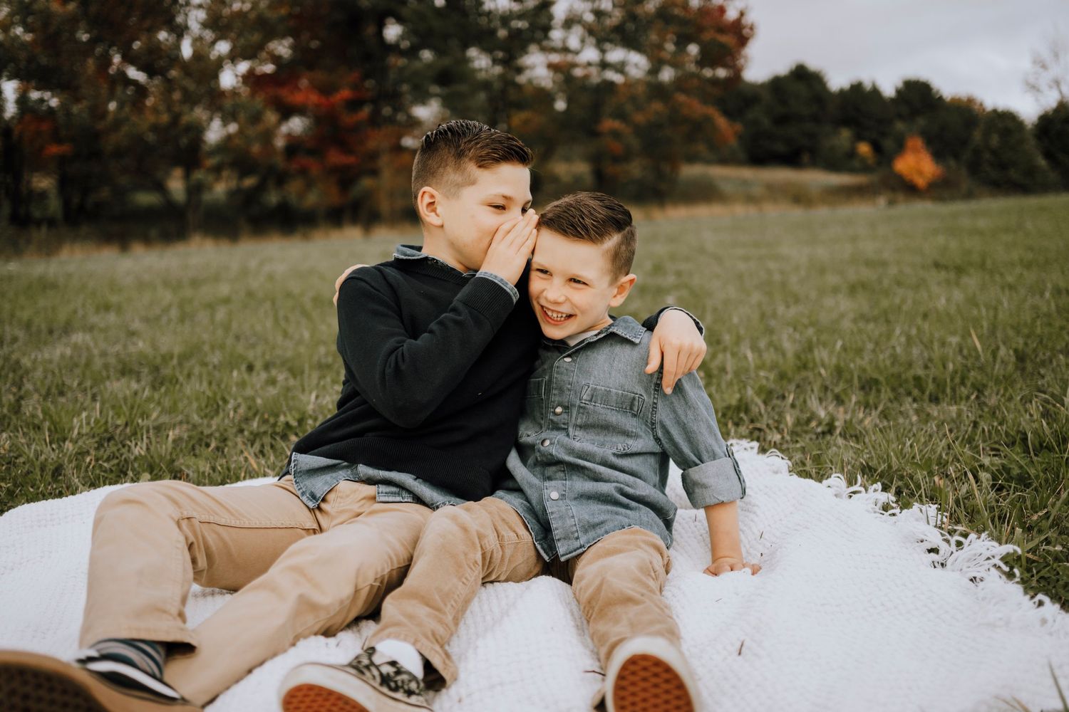 Two siblings sharing a sweet moment while sitting together on a white blanket in a grassy field.