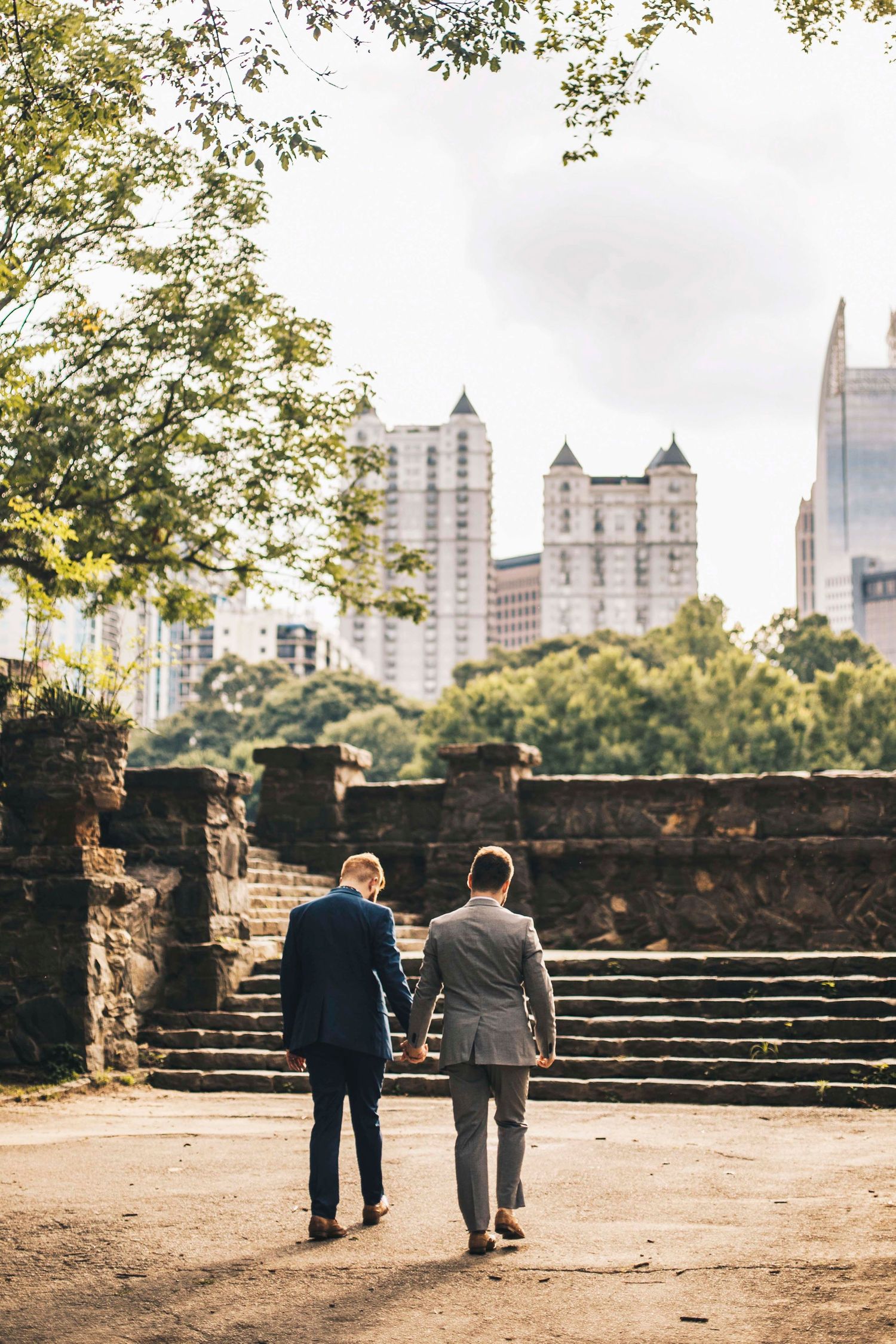 Two men in suits hold hands while walking through an urban park with city skyline in background.