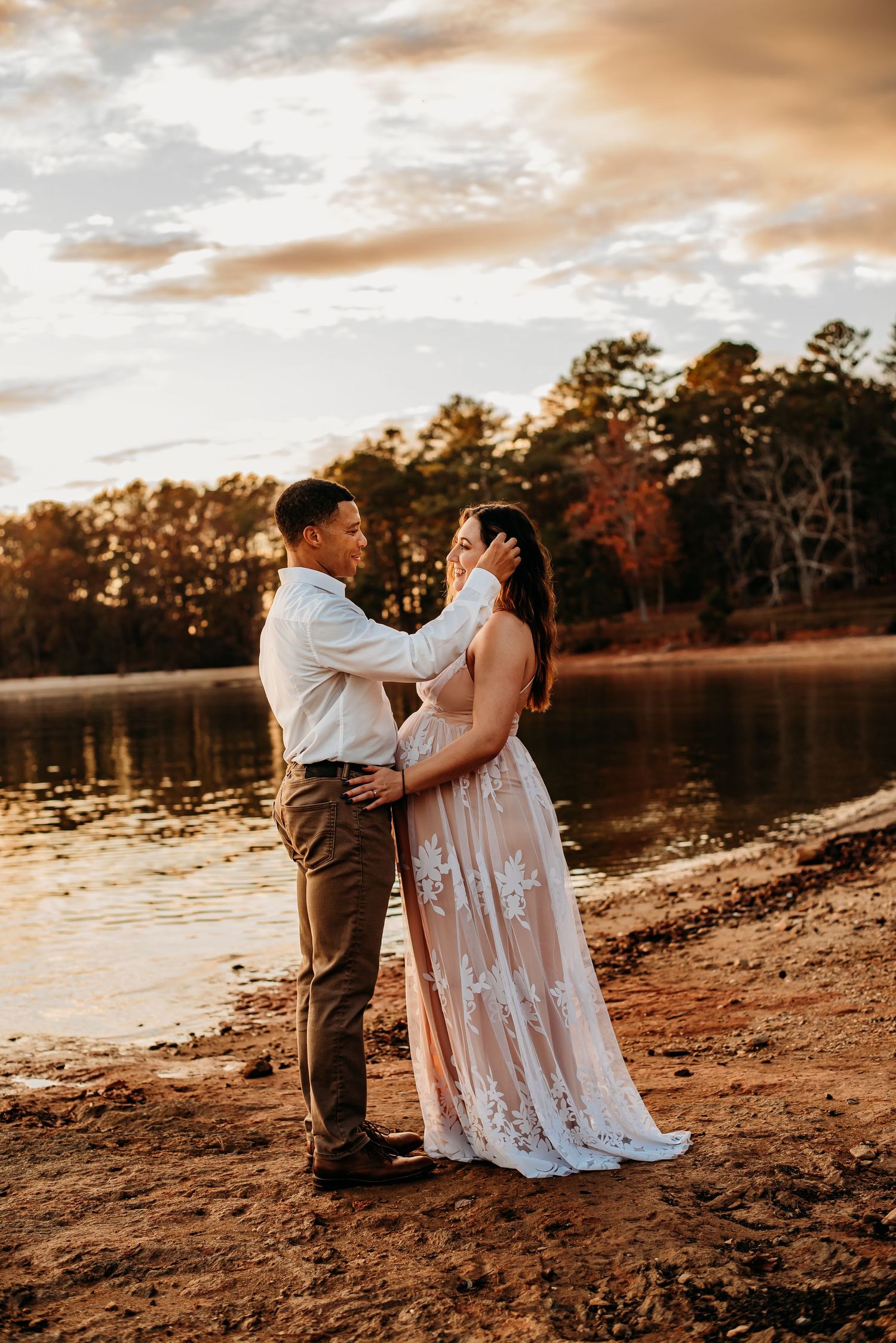 Couple embraces romantically at sunset by a lake, woman wearing an elegant lace wedding gown.