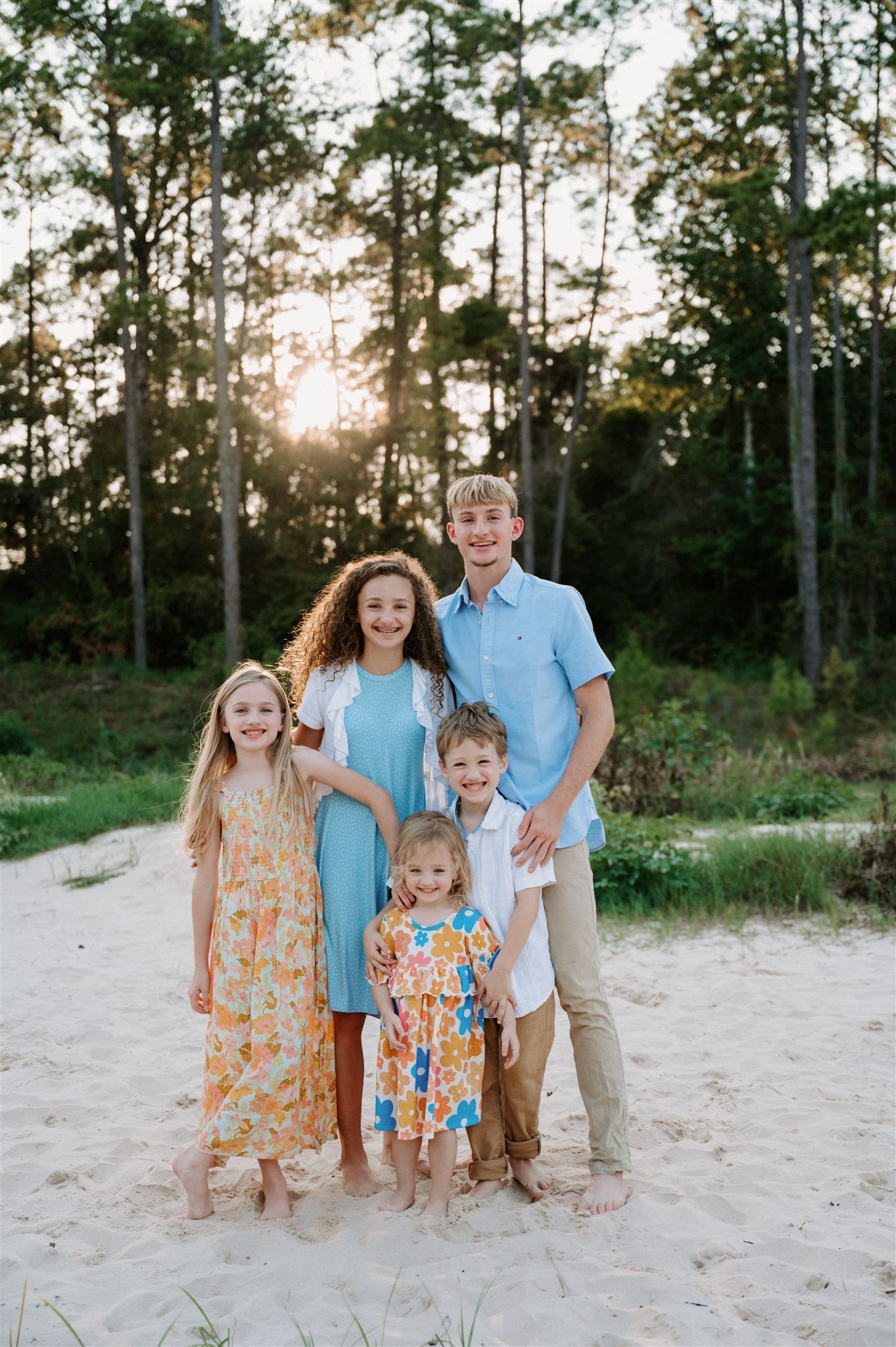 Family group standing together on sandy beach path at sunset with tall pine trees in background.