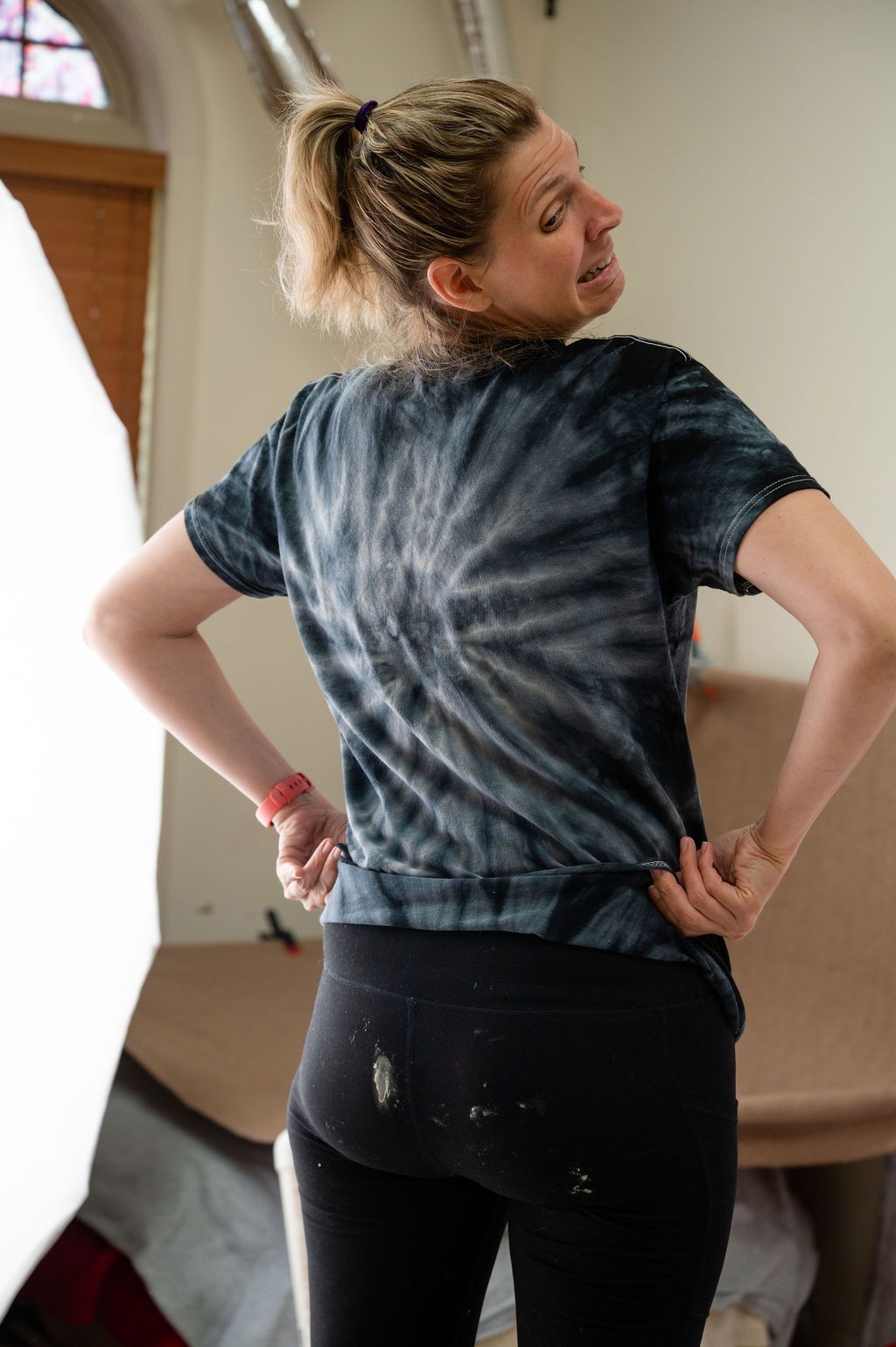 A person wearing a tie-dye navy t-shirt and black leggings stands with hands on hips in a room with arched windows.