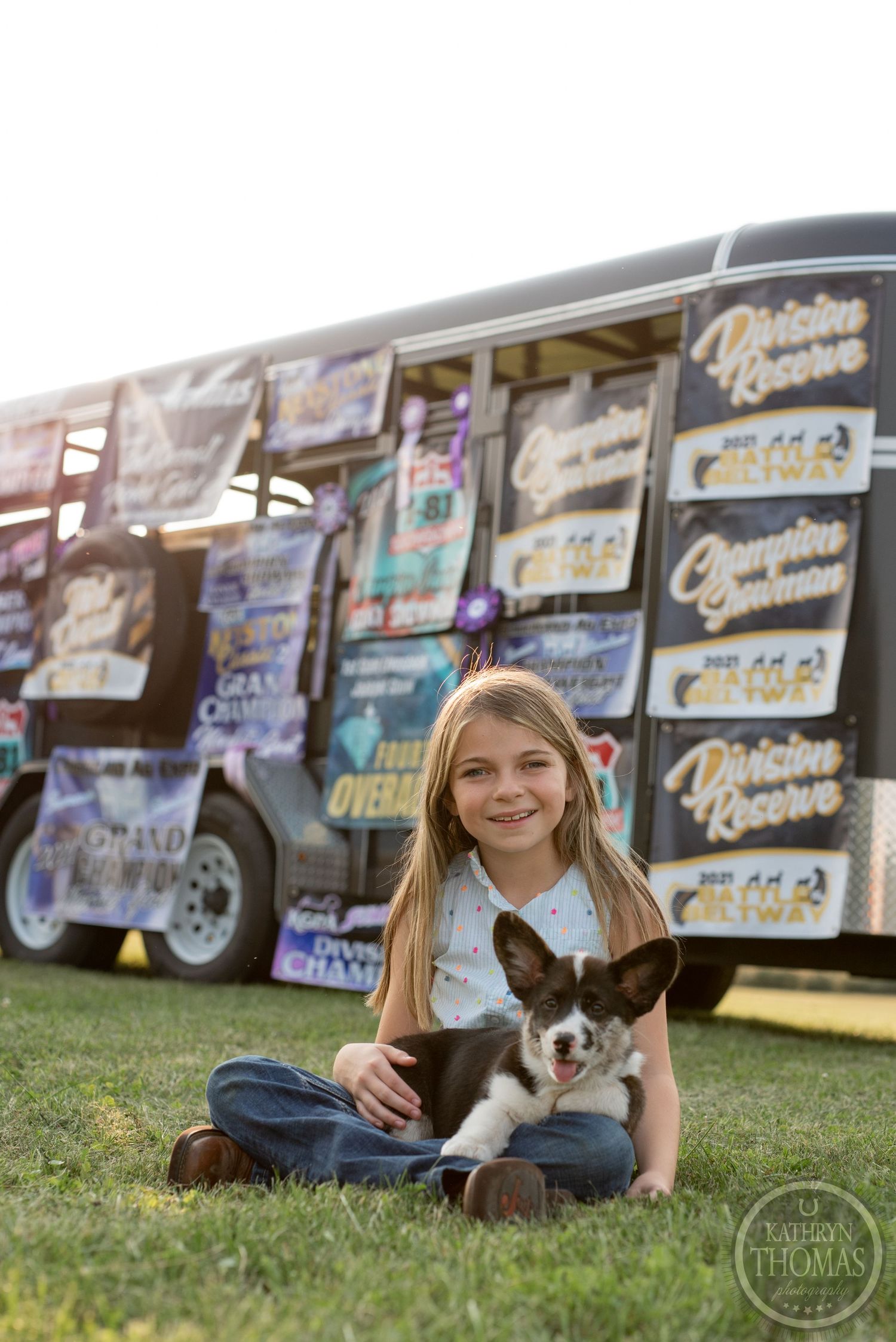 Person sits with small dog on grass in front of wall of show banners.