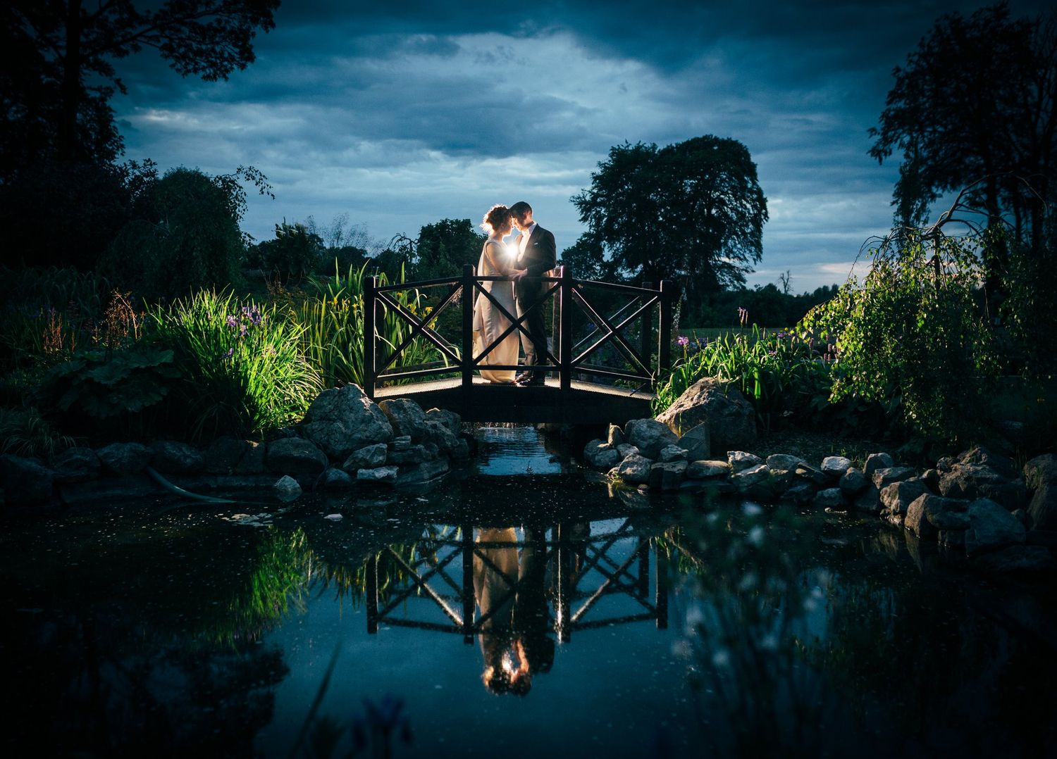 Dramatic blue twilight landscape with bridge silhouette reflected in still water.