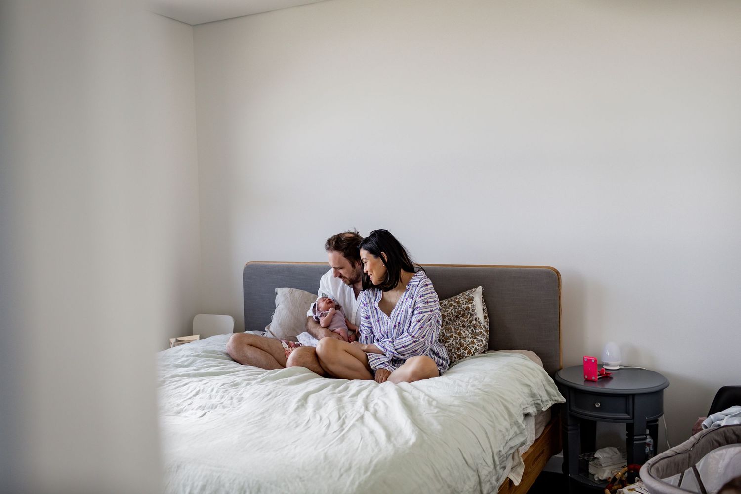 Couple sitting on grey bed against white wall in casual home setting.