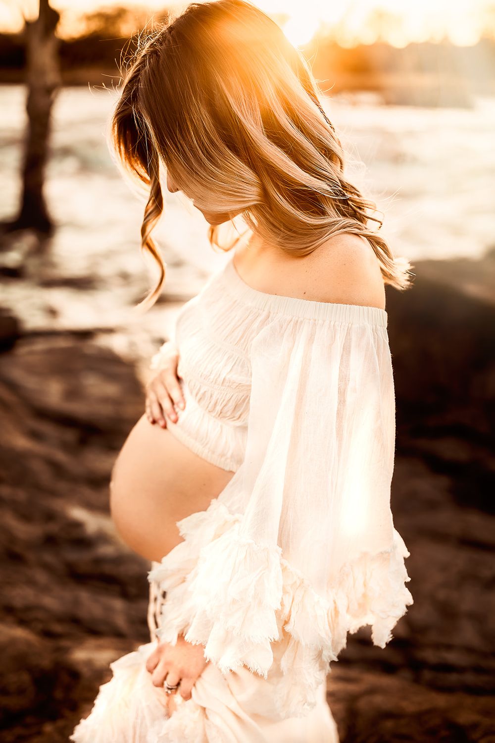 A pregnant mother holds her belly while the sun shines behind her during golden hour.