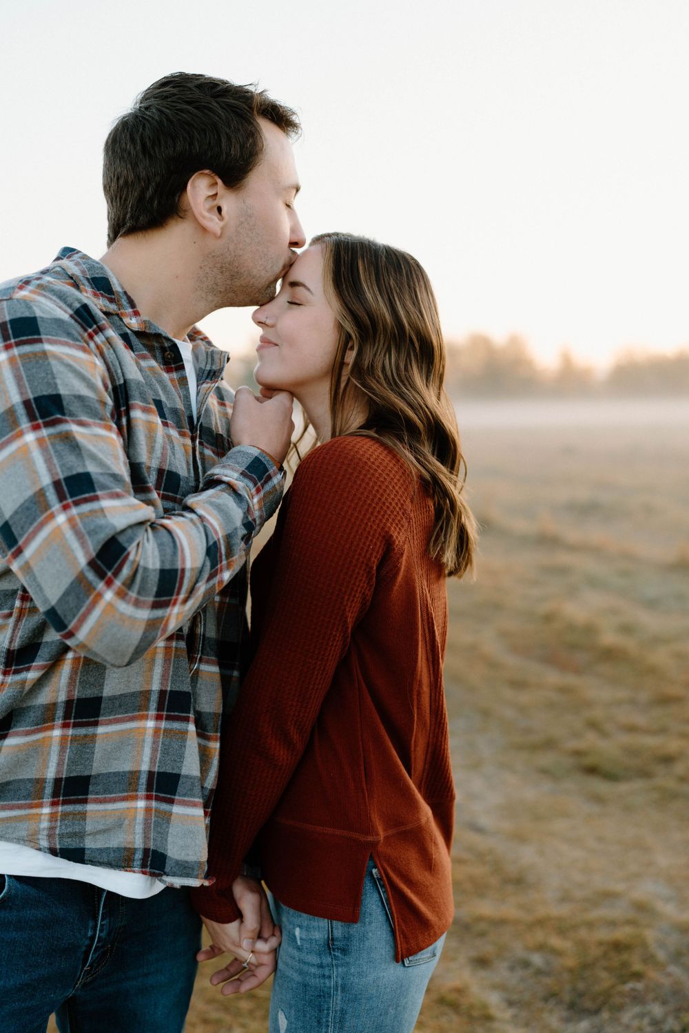 A couple in plaid and burgundy shares tender moments in a field during sunset.